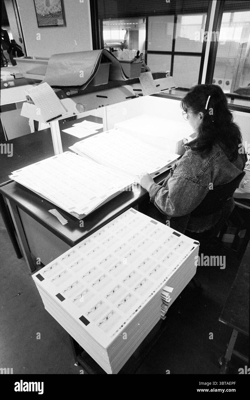 Società di stampa varie Enschedéen Zn. Enschede & Zn industria grafica MV Joh. Haarlem Paesi Bassi, Whizgle News, Dutch Desk, Paesi Bassi, 1950 - 2000 il 26-04-1988. L'immagine mostra questi argomenti. In un ambiente interno spazioso, una donna è seduta a una scrivania, molto concentrata su un compito. Ha lunghi capelli scuri che si arrampicano sulle spalle, leggermente oscurati dal panino che è fissato con una matita. Indossando una giacca di denim, si piega in avanti, le braccia appoggiate sul tavolo, mentre esamina una grande pila di carta che giace davanti a lei. La scrivania è piena di fogli, ognuno dei quali mostra un Foto Stock