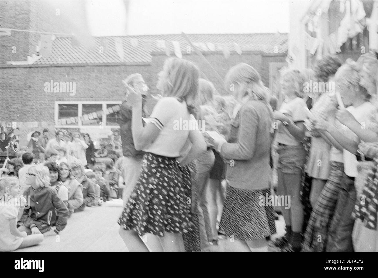 Performance dei bambini + mercato in occasione del 50° anniversario degli anniversari giubilari della scuola Willem de Zwijger, Whizgle News, Dutch Desk, Paesi Bassi, 1950 - 2000 il 22-06-1974. L'immagine mostra questi argomenti. La scena presenta un vivace raduno all'aperto, probabilmente un evento scolastico o comunitario. In primo piano, un gruppo di bambini sembra ballare e divertirsi. Indossano una varietà di abbigliamento casual tipico della cultura giovanile, tra cui gonne corte, T-shirt e abiti a fantasia, suggerendo un'atmosfera spensierata. I colori della scena sono un mix di toni chiari Foto Stock