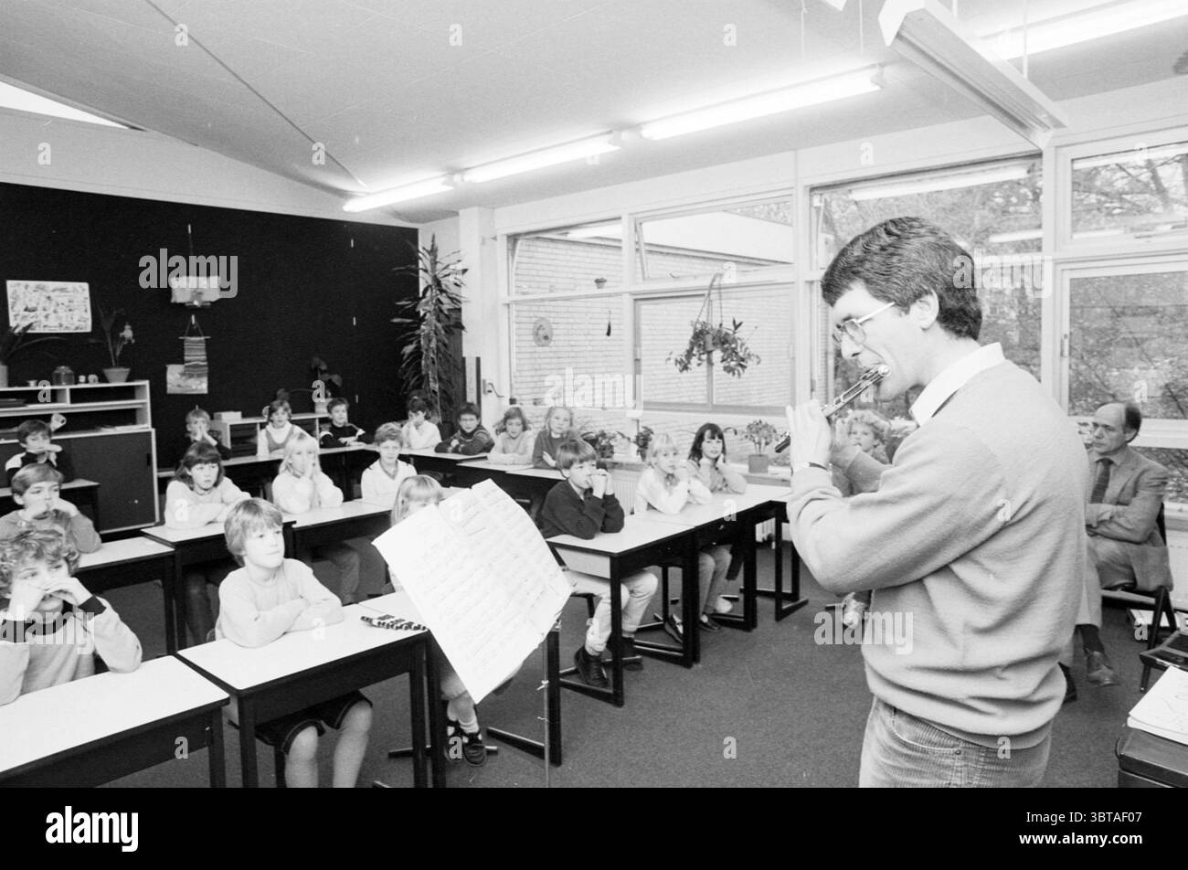 Flautista NPO insegna alla scuola Driehuis North Holland Philharmonic Orchestra Schools Driehuis, Whizgle News, Dutch Desk, Paesi Bassi, 1950 - 2000 il 21-11-1984. Questi sono gli elementi dell'immagine. La scena è ambientata in un'aula dall'atmosfera calda e invitante. Le pareti sono per lo più di tonalità neutre, incluso uno sfondo scuro che contrasta con le luminose e ariose finestre. La luce naturale si filtra, proiettando ombre morbide e creando una luminosità delicata che riempie lo spazio. Davanti alla stanza, un insegnante si erge, gestendo animatamente con le mani. Trasuda entusiasmo mentre si impegna Foto Stock
