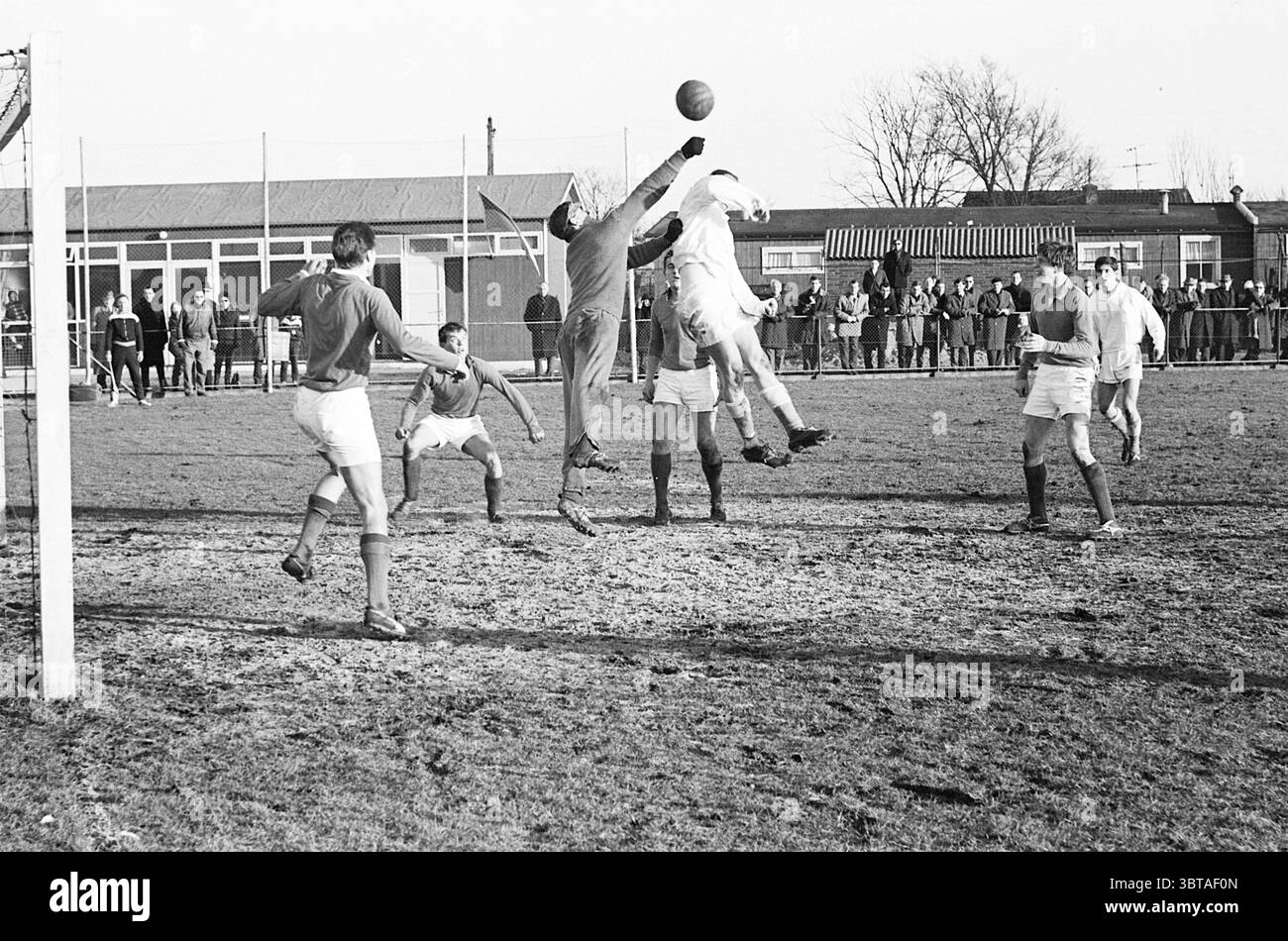 E.H.S. - D.C.O. Football, Whizgle News, Dutch Desk, Paesi Bassi, 1950 - 2000 il 31-01-1965. Questi sono gli argomenti nell'immagine. Nella scena, una partita di calcio dinamica si svolge su un campo erboso ben consumato, caratterizzato da macchie di terra marrone e grumi d'erba. In primo piano, diversi giocatori sono impegnati in un momento di azione intensa. Un giocatore, rivestito di maglia a maniche lunghe e pantaloncini corti, salta in aria, braccia allungate, tentando di catturare la palla sopra compagni di squadra e avversari. La sua espressione focalizzata mostra determinazione e determinazione. Intorno a lui, altri giocatori sono disposti in varie pose - s Foto Stock
