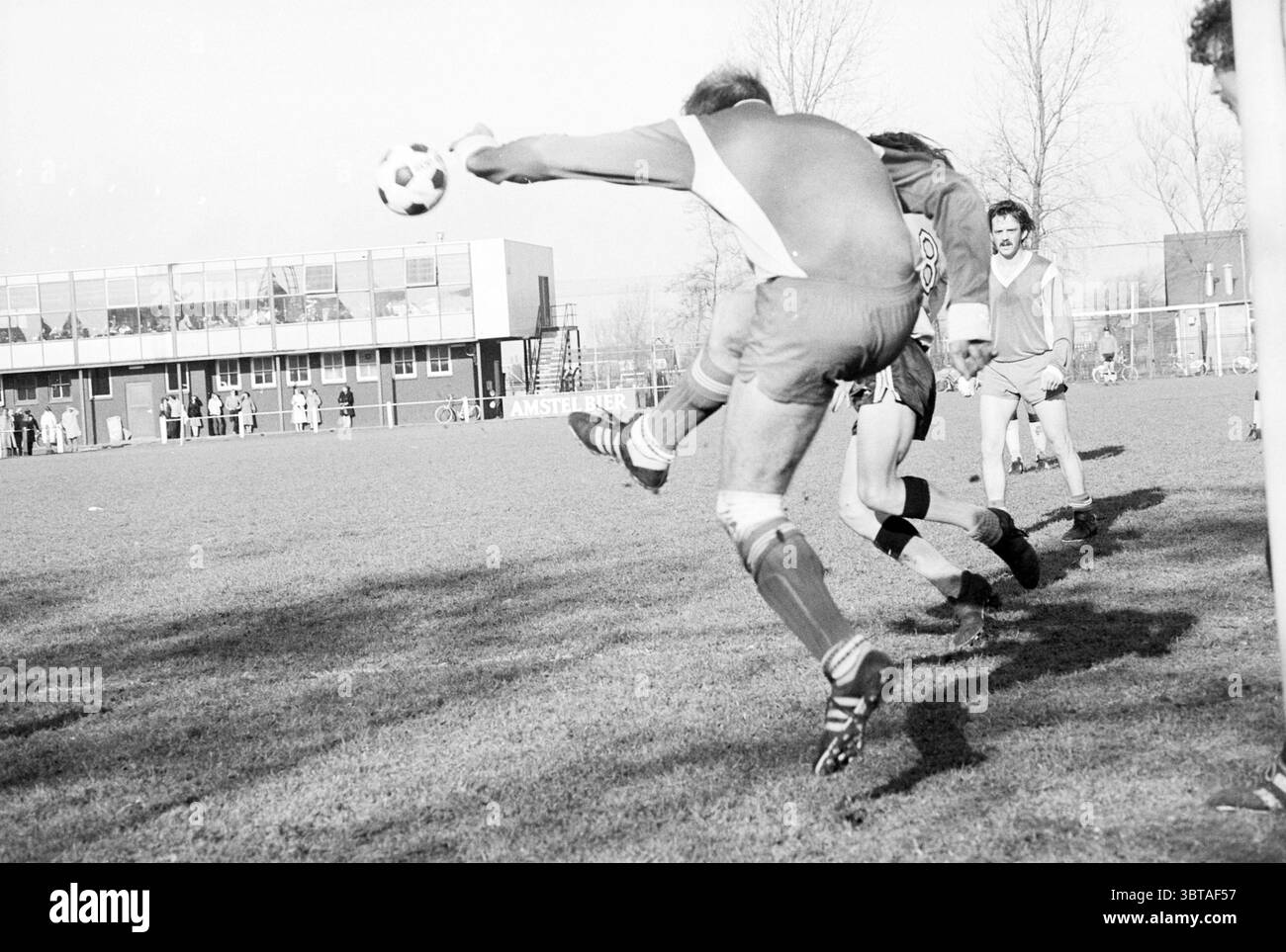 Schoten - The Bridge Football, Whizgle News, Dutch Desk, Paesi Bassi, 1950 - 2000 il 10-02-1980. Questi sono gli argomenti nell'immagine. La scena cattura un momento dinamico su un campo da calcio, dove un giocatore con una maglia rossa viene trasportato in aria, colpendo abilmente una palla da calcio con il piede. L'espressione determinata del giocatore suggerisce un'intensa concentrazione sul gioco. Intorno a loro, compagni di squadra e avversari mostrano un mix di impegno e tensione, con alcuni giocatori pronti a rispondere al gioco. Il campo stesso è rigoglioso e verde, in contrasto con la nitidezza delle uniformi dei giocatori, tra cui b Foto Stock