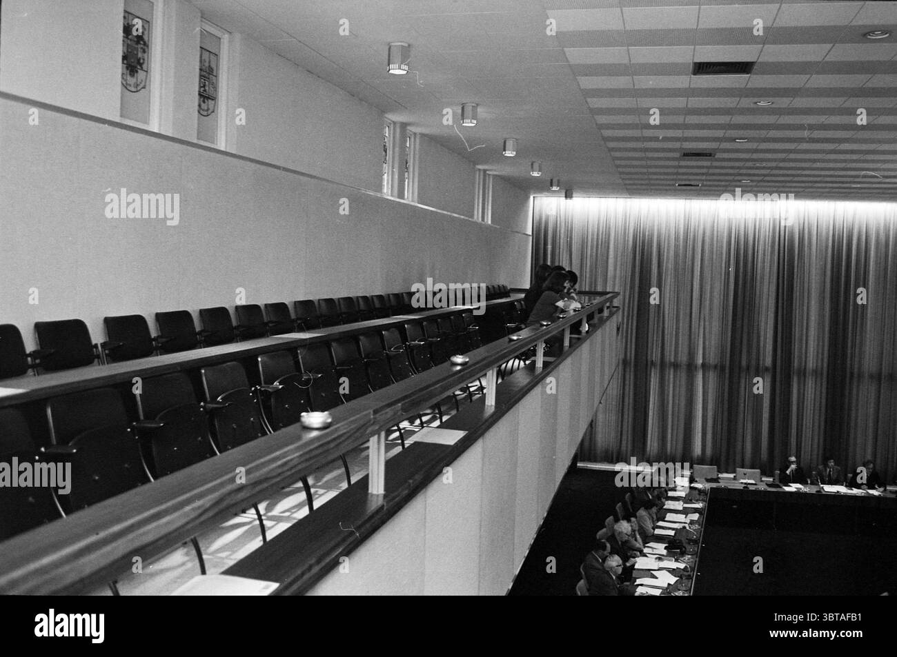 Meeting Social Workers Meetings, Whizgle News, Dutch Desk, Paesi Bassi, 1950 - 2000 il 24-05-1973. Questi argomenti vengono visualizzati nell'immagine. La scena raffigura un interno spazioso, probabilmente una grande sala riunioni o conferenze. L'ambientazione è caratterizzata da un soffitto alto con una griglia di lampade che illuminano delicatamente l'area. La tavolozza dei colori è silenziata, basandosi su sfumature di grigio e nero, il che suggerisce un'atmosfera seria. In primo piano, una lunga ringhiera di legno separa il livello superiore della sede dal livello inferiore. Le più file di sedie nere sono disposte in modo ordinato, creando ombre morbide sul pavimento Foto Stock