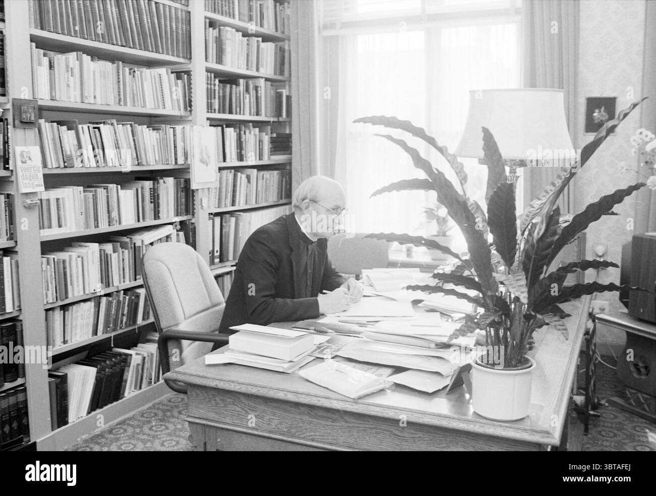 Bishop Bomers of Haarlem Roman Catholic Church Haarlem Paesi Bassi, Whizgle News, Dutch Desk, Paesi Bassi, 1950 - 2000 il 03-09-1984. Questi sono gli elementi dell'immagine. In un ambiente interno pieno di calore di luce naturale che filtra attraverso le grandi finestre, un uomo anziano siede su una scrivania in quercia, profondamente coinvolto nel suo lavoro. Indossa un vestito oscuro e formale, che suggerisce un comportamento accademico. I suoi capelli sono grigi e spessi, e sembra essere in una concentrazione ponderata, con una mano che tiene una matita su fogli di carta sparsi per la scrivania. Intorno a lui, una ricca dimostrazione di Foto Stock