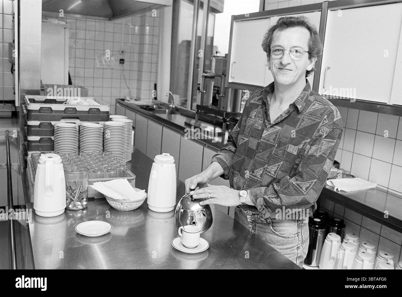 WLTO Workplaces, Whizgle News, Dutch Desk, Paesi Bassi, 1950 - 2000 il 18-12-1994. L'immagine mostra questi argomenti. La scena cattura una cucina luminosa e ariosa, piena di calore e attività. I ripiani in acciaio inox riflettono una luce soffusa, conferendo allo spazio un aspetto pulito e moderno. Dietro la figura centrale, le alte finestre consentono alla luce naturale di inondare, illuminando vari utensili da cucina ed elettrodomestici allineati in modo ordinato contro le pareti. In primo piano, una persona con capelli ricci si trova accanto a un distributore di bevande in metallo lucido, versando una bevanda calda in una tazza bianca appoggiata su una sa Foto Stock