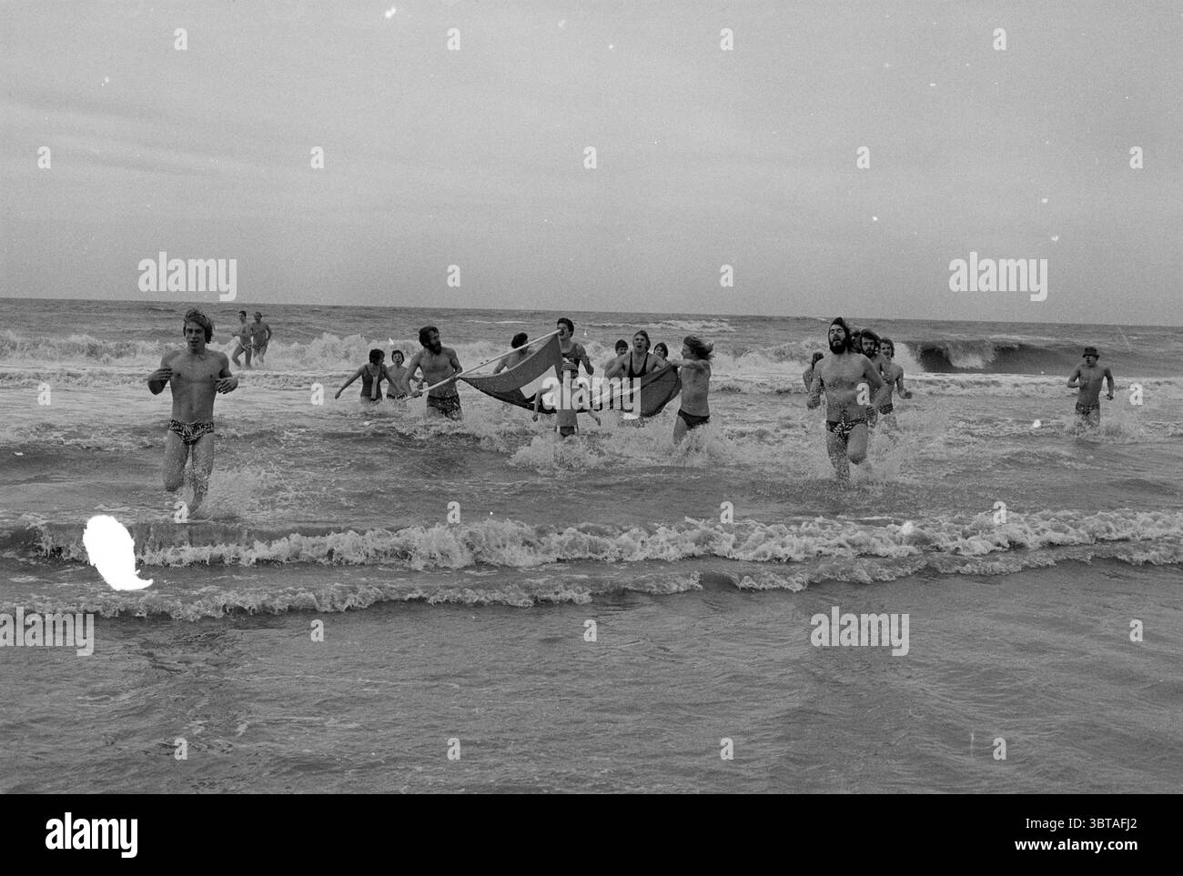 Immersione di Capodanno nel mare di DER Capodanno, Whizgle News, Dutch Desk, Paesi Bassi, 1950 - 2000 il 01-01-1977. Questi sono gli elementi dell'immagine. Una vivace scena sul lungomare si dispiega, con un gruppo di persone che spruzzano attraverso le onde poco profonde dell'oceano. L'atmosfera è energizzata, piena di risate e senso di libertà. Gli individui, rivestiti di costumi da bagno, mostrano un mix di divertimento e allegria spensierata mentre corrono verso l'acqua. Le loro espressioni sono vivaci e mostrano una serie di emozioni, dall'euforia alla pura gioia. La riva è punteggiata di sabbia soffice e dorata Foto Stock