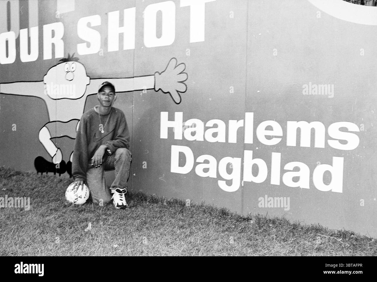 Vincitore Shoot Your Shot 'Lloyd' C Cup Haarlem [Toneelschuur Youth film competition Coca Cola Haarlem Cup] Haarlem Paesi Bassi, Whizgle News, Dutch Desk, Paesi Bassi, 1950 - 2000 il 29-07-1997. Questi argomenti vengono visualizzati nell'immagine. In un ambiente urbano dinamico, un giovane si inginocchia su un'erba, indossando un abbigliamento rilassato che suggerisce comfort e casualità. Il soggetto ha un'espressione amichevole, trasuda un senso di orgoglio e fiducia. Dietro di loro c'è un muro vibrante ricoperto da opere d'arte in stile graffiti, con un personaggio cartoni animati con un grande e esagerato reac a mano Foto Stock