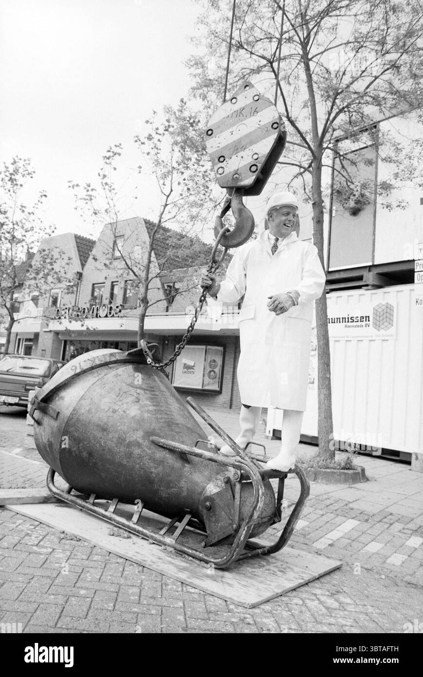 Punto più alto del pozzo a Heemstede Heemstede Nederland, Whizgle News, Dutch Desk, Paesi Bassi, 1950 - 2000 il 19-06-1991. L'immagine include questi argomenti. La scena vede un uomo in piedi con sicurezza su un grande oggetto dall'aspetto industriale che assomiglia a un pezzo fuso pesante o a un meccanismo di sollevamento. Indossa un lungo cappotto bianco, che contrasta nettamente con gli elementi circostanti. La sua espressione è di orgoglio o di divertimento, aggiungendo un senso di leggerezza all'ambiente altrimenti robusto. Sullo sfondo, ci sono modesti edifici a due piani con tetti inclinati e grandi finestre, a suggerire Foto Stock