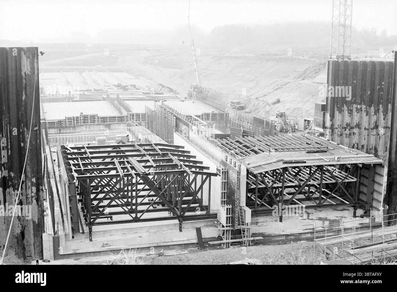 Construction Wijkertunnel Velsen Velsen, Whizgle News, Dutch Desk, Paesi Bassi, 1950 - 2000 on 28-10-1993. Questi sono gli argomenti nell'immagine. La scena raffigura un ampio cantiere caratterizzato da una tavolozza di colori tenui di grigi e neri, che suggerisce un ambiente di lavoro dominato da acciaio e cemento. In primo piano, si innalza un grande telaio in acciaio, le sue linee angolari formano una struttura geometrica che indica un progetto ingegneristico significativo. Numerose gru sono visibili in lontananza, con le loro silhouette affilate contro i cieli nuvolosi, accentuando un senso di altezza e AC Foto Stock