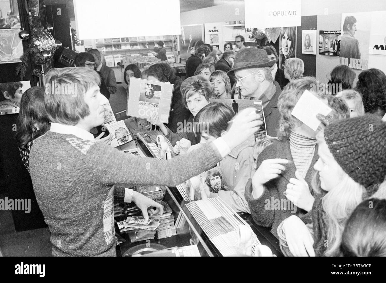 Udo Jurgens at Mic Music, Whizgle News, Dutch Desk, Paesi Bassi, 1950 - 2000 il 21-12-1971. Questi argomenti vengono visualizzati nell'immagine. La scena cattura un ambiente interno vivace, probabilmente un negozio di dischi o un negozio simile. Il primo piano presenta un bancone circondato da un gruppo diversificato di persone. Una persona, forse un cassiere o un impiegato, è attivamente coinvolta con i clienti, gestendo con una mano tenendo una pila di documenti nell'altra. Il loro abbigliamento suggerisce uno stile retrò, con un maglione che aggiunge un tocco di calore alla scena. Intorno a loro, una folla di acquirenti entusiasti mostra una V Foto Stock