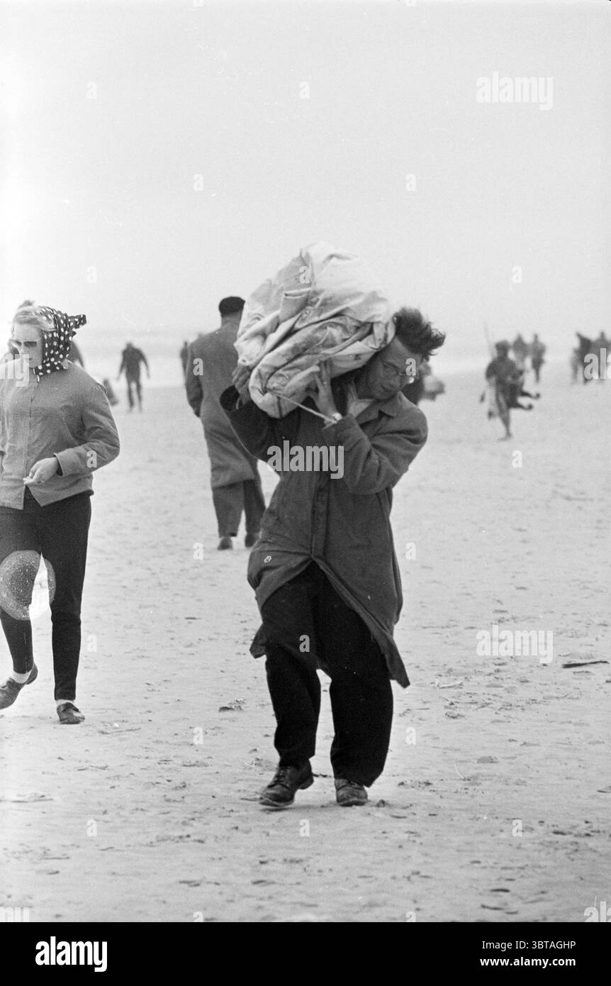 Fishermen on the Beach Beach and Beaches Fisheries and other fishmongers fishing, Whizgle News, Dutch Desk, Paesi Bassi, 1950 - 2000 il 14-01-1963. Questi sono gli argomenti nell'immagine. In questa scena costiera, i toni morbidi e silenziosi del grigio e del bianco dominano l'atmosfera, creando un senso di freschezza e luce soffusa. La spiaggia si estende, sabbiosa e piatta, con dolci onde che si infrangono sullo sfondo. Un uomo è al centro della scena, strisciando intenzionalmente sulla sabbia. Porta un grande oggetto impacchettato sulla sua spalla, stretto in tessuto, suggerendo un'aria di determinazione e di sforzo. H Foto Stock