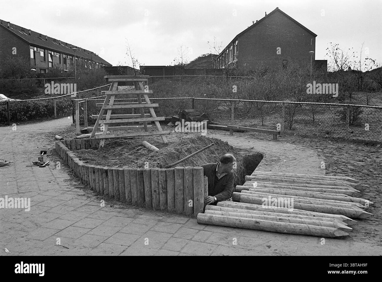 Costruzione di attrezzature da gioco IJmuiden Playgrounds and Playgrounds, Whizgle News, Dutch Desk, Paesi Bassi, 1950 - 2000 su 15-11-1976. Questi argomenti sono mostrati nell'immagine. La scena è un parco giochi che combina elementi naturali e aree di gioco costruite, realizzate in tonalità monocromatiche, che evocano un'atmosfera nostalgica o storica. In primo piano, c'è una scatola di sabbia a forma di cerchio ampio, con una barriera di legno che la circonda. Un bambino si inginocchia accanto a questa zona circolare, intensamente impegnato nel gioco. La forma del bambino suggerisce una concentrazione profonda, magari scavando o creando qualcosa Foto Stock