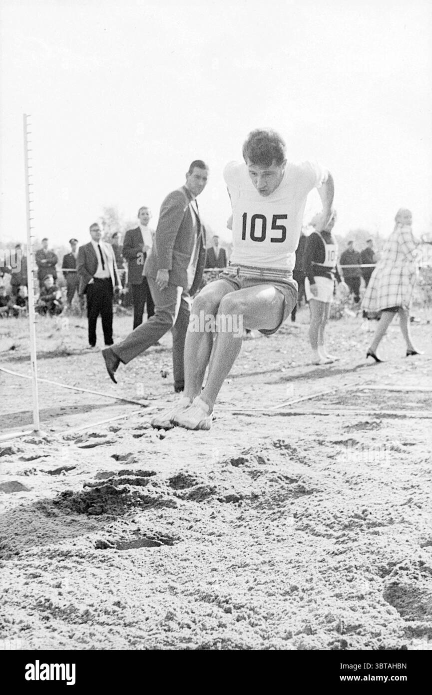 6 stelle dello sport provi Decathlon Sports and Sports Week, Whizgle News, Dutch Desk, Paesi Bassi, 1950 - 2000 il 15-10-1968. Questi argomenti vengono visualizzati nell'immagine. La scena cattura un momento dinamico in una gara di salto lungo. In primo piano, un atleta, concentrato e determinato, viene catturato a mezz'aria, sospeso sopra un pozzo di atterraggio sabbioso. Indossa una maglia bianca aderente con il numero "105" in evidenza, insieme a pantaloncini sportivi abbinati. Le sue gambe muscolari sono allungate in avanti, mostrando forma atletica, mentre le sue braccia sono posizionate per l'equilibrio. Sullo sfondo, una folla sparsa osserva Foto Stock
