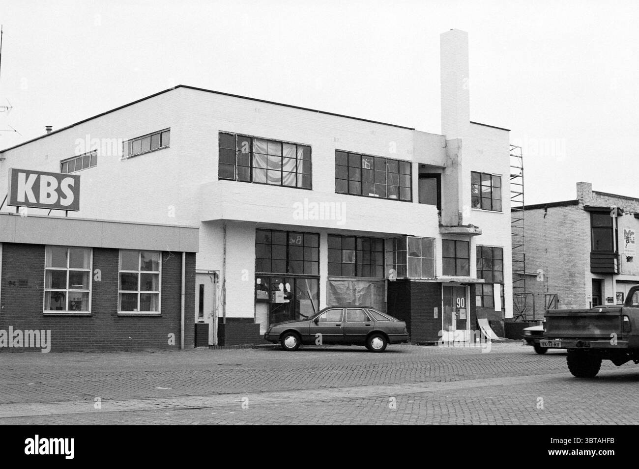 Trawlerkade 90 IJmuiden IJmuiden Trawlerkade Paesi Bassi, Whizgle News, Dutch Desk, Paesi Bassi, 1950 - 2000 il 16-01-1992. Questi sono gli elementi dell'immagine. La scena presenta un edificio industriale della metà del secolo caratterizzato dalle sue linee pulite e dal design funzionale. La struttura presenta una facciata prevalentemente bianca, che contrasta nettamente con gli elementi circostanti. Grandi finestre, incorniciate in nero, dominano la parte anteriore, consentendo scorci dell'interno. Le finestre hanno un'atmosfera industriale, spesso vista in ambienti commerciali. In primo piano, c'è una superficie di mattoni, probabilmente un parcheggio Foto Stock