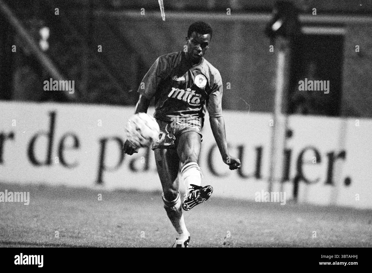 Calcio: Haarlem - Emmen Emmen, Whizgle News, Dutch Desk, Paesi Bassi, 1950 - 2000 il 26-10-1990. L'immagine contiene questi argomenti. La scena cattura un momento dinamico in un evento sportivo, probabilmente una partita di calcio. Un atleta maschile è presente in primo piano, a metà azione mentre colpisce un pallone da calcio con un'intensa concentrazione. La sua montatura atletica suggerisce che è altamente abile, mostrando una combinazione di potenza e precisione nella sua postura. Indossa una maglia scura impreziosita da un emblema di colore chiaro, che suggerisce l'affiliazione di una squadra. Il colore della maglia contrasta nettamente con la luminosità dell'ambiente, dra Foto Stock