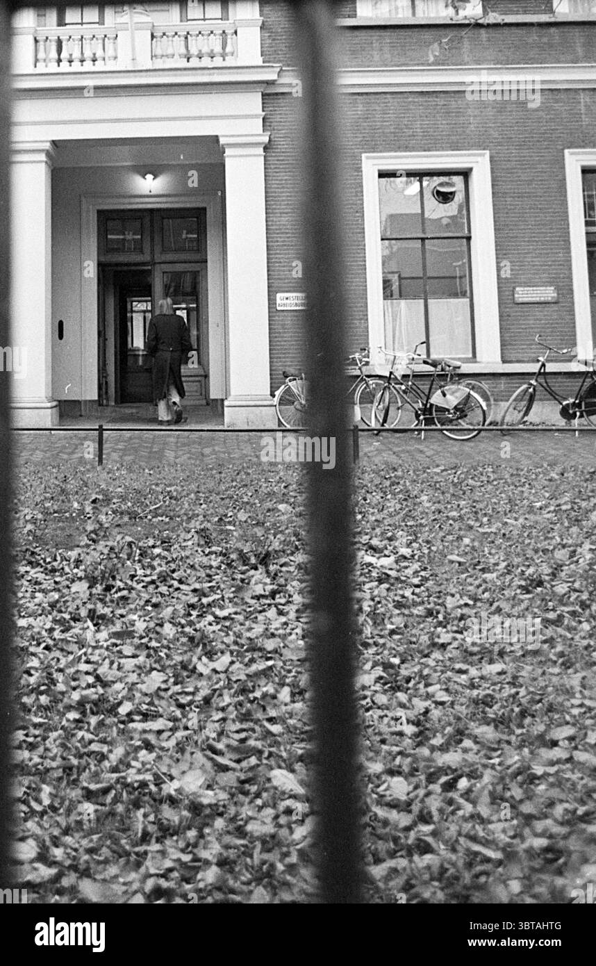 Ufficio regionale del lavoro Haarlem Paesi Bassi., Whizgle News, Dutch Desk, Paesi Bassi, 1950 - 2000. Questi sono gli argomenti nell'immagine. In questa scena, l'attenzione è rivolta all'ingresso di un edificio incorniciato da sbarre, suggerendo un senso di separazione o confinamento. La struttura presenta un design classico con una facciata robusta, caratterizzata dalla sua muratura e dall'entrata prominente. Si vede una figura che cammina attraverso l'ingresso, vestita con abiti scuri, aggiungendo un senso di movimento alla quiete dell'ambiente circostante. Sparse per tutto il primo piano sono cadute foglie, suggerendo la stagione e c Foto Stock
