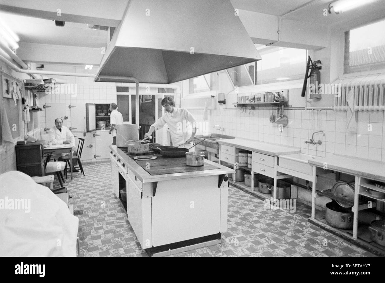 Cucina istituzionale con tre dipendenti, Whizgle News, Dutch Desk, Paesi Bassi, 1950 - 2000 il 01-02-1979. L'immagine include questi argomenti. La scena è ambientata in una spaziosa cucina, caratterizzata da una tavolozza prevalentemente monocromatica, che le conferisce un aspetto pulito e funzionale. Le pareti sono piastrellate in bianco, creando un ambiente luminoso e sterile, mentre il pavimento a scacchi grigio e bianco aggiunge un tocco di interesse visivo. In primo piano spicca una grande isola di cucina centrale. E' dotata di un'ampia fornelli e di un cofano metallico sopra il tetto, da cui alcuni tubi lucidi Foto Stock