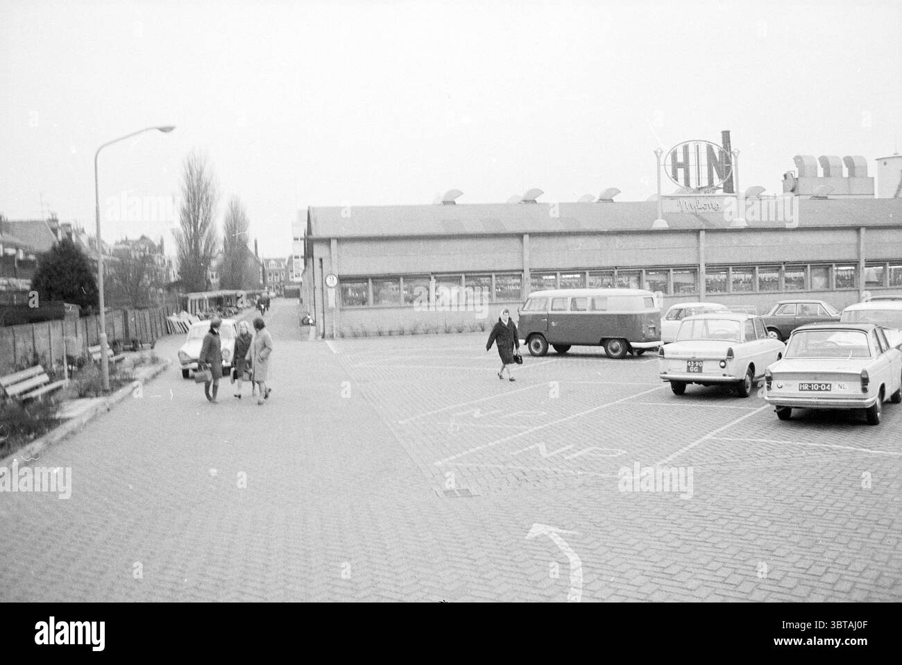Est. HIN Exterior, Whizgle News, Dutch Desk, Paesi Bassi, 1950 - 2000 il 05-02-1969. Questi sono gli argomenti nell'immagine. La scena cattura un parcheggio semi-urbano, prevalentemente monocromatico, che offre un'atmosfera nostalgica. Un gruppo di tre donne vestite con cappotti della metà del secolo passeggiano verso il lato sinistro dello spazio, mostrando gli stili tipici dell'epoca con le loro varie silhouette. Le donne appaiono impegnate nella conversazione, contribuendo al senso di vita e al dinamismo dell'ambiente altrimenti tranquillo. In primo piano, la superficie pavimentata del parcheggio è fiancheggiata da posti auto, S Foto Stock