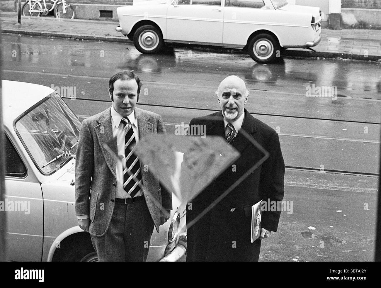 Gestione di Moppes Diamantairs in Amsterdam Persons, Whizgle News, Dutch Desk, Paesi Bassi, 1950 - 2000 il 22-10-1974. L'immagine include questi argomenti. In una scena di strada caratterizzata da una tavolozza di colori tenui, due uomini stanno fianco a fianco, osservando con un senso dello scopo. Il primo uomo, che indossa un abito grigio chiaro con cravatta a righe, ha un aspetto leggermente formale, mentre il secondo uomo è vestito con un soprabito scuro, suggerendo uno stile più classico. Le loro espressioni sono contemplative, come se fossero impegnate in conversazione o riflettendo su qualcosa di importante. Lo sfondo rivela una giornata di pioggia; t Foto Stock