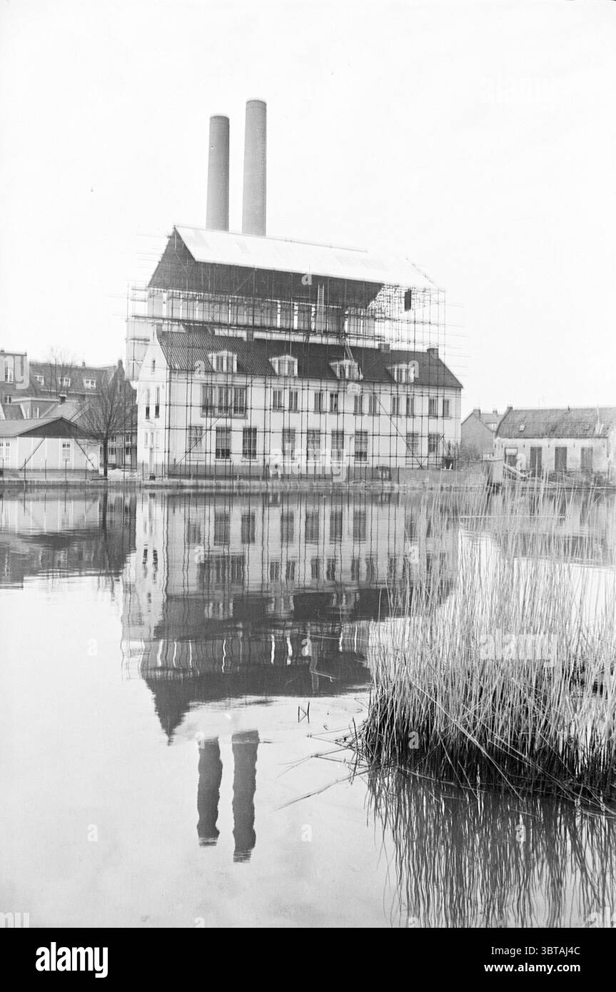 Piccolo padiglione della Fondazione Maria in case impalcate e costruzione di case, Whizgle News, Dutch Desk, Paesi Bassi, 1950 - 2000 il 15-04-1964. Questi argomenti vengono visualizzati nell'immagine. La scena presenta un grande edificio industriale, caratterizzato dalla sua architettura distintiva. La struttura si erge in modo prominente lungo il bordo di un corpo d'acqua, il cui riflesso si specchia sulla superficie calma. Due alte aree fumatori sorgono dal tetto, che creano ombre allungate sull'area circostante. L'edificio stesso e' avvolto da impalcature, suggerendo che sia in fase di ristrutturazione o costruzione. L'esterno Foto Stock