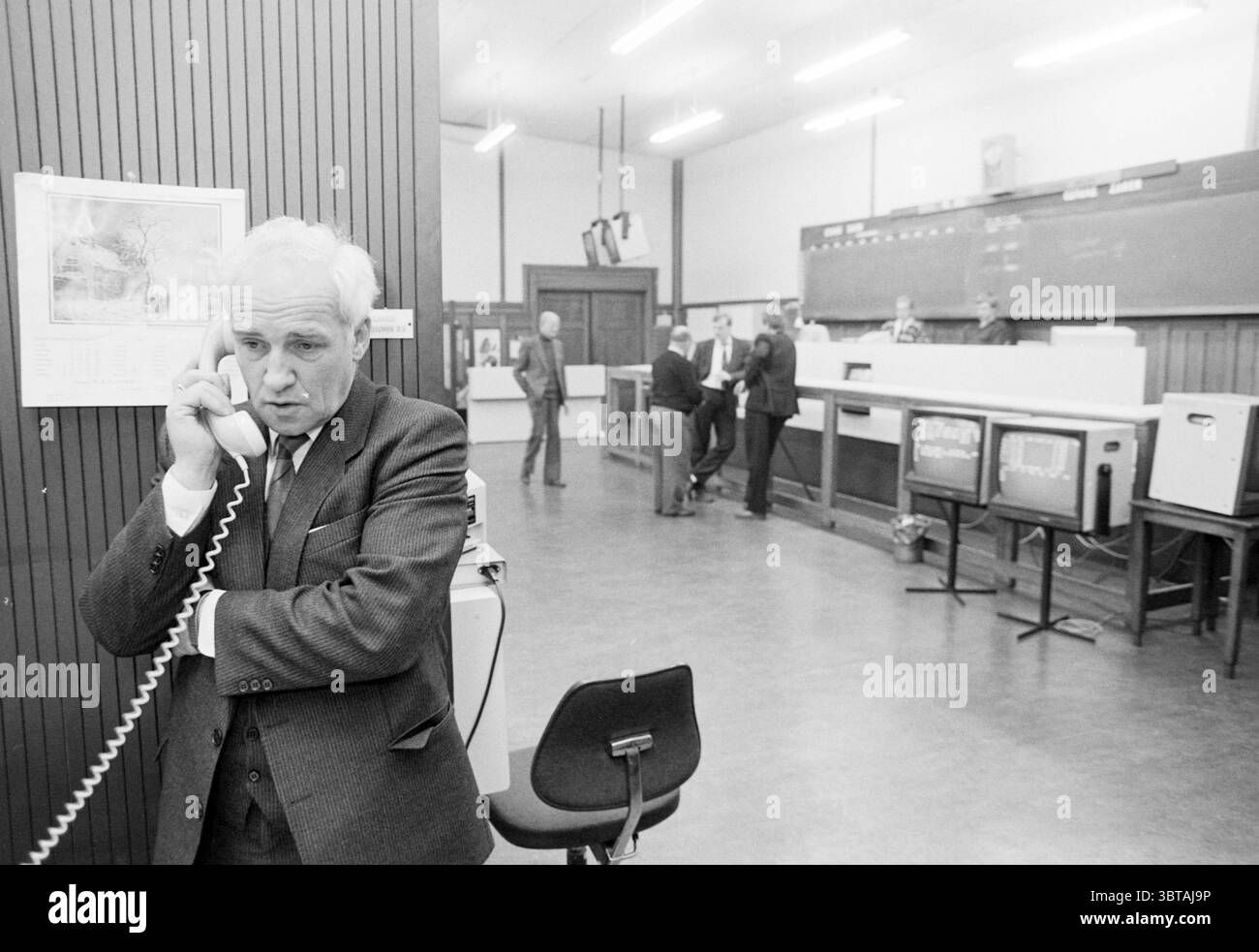 Sig. Nieuwenhuizen potato futures market mercante fiera orticoltura, Whizgle News, Dutch Desk, Paesi Bassi, 1950 - 2000 il 21-01-1987. Questi argomenti sono mostrati nell'immagine. La scena raffigura un ambiente interno affollato, che ricorda un ufficio o una sala di controllo della metà del XX secolo. Un uomo si trova in primo piano, impegnato in una conversazione telefonica. Ha un'espressione seria, vestito con un abito scuro con una camicia di colore chiaro, e si appoggia contro un divisore o un muro. La sua postura suggerisce un misto di urgenza e contemplazione, con l'altro braccio attraversato sul petto. Sullo sfondo, sever Foto Stock