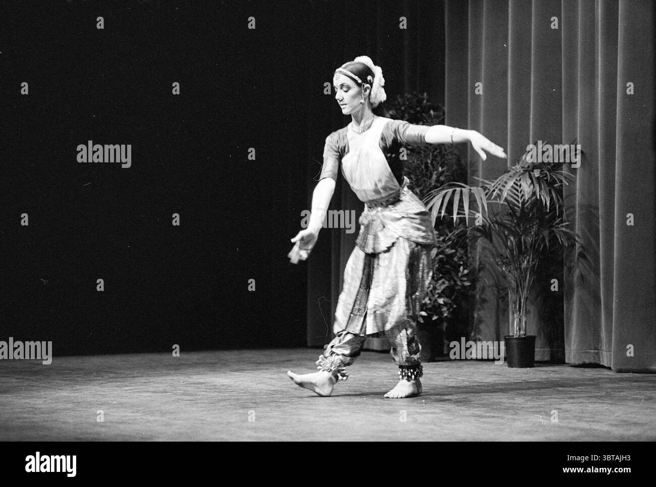 Danze indiane Nieuwe Slof B'wijk Dancing, Whizgle News, Dutch Desk, Paesi Bassi, 1950 - 2000 il 12-03-1977. L'immagine mostra questi argomenti. La scena cattura una ballerina a metà spettacolo, posizionata con grazia su un palcoscenico scarsamente illuminato, sottolineando l'artista. È adornata con abiti tradizionali caratterizzati da tessuti vivaci e fantasie intricate che esaltano i suoi movimenti. La ballerina indossa un top aderente e una gonna fluida che oscilla leggermente mentre si muove, con accessori elaborati che catturano l'attenzione. I suoi arti si estendono fluidamente, trasmettendo forza ed eleganza. Il de Foto Stock