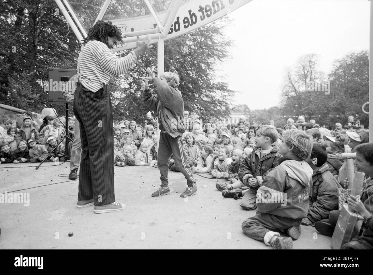 Panettieri da festa Haarlemmerhout esibizione di Hakim Traidia, Whizgle News, Dutch Desk, Paesi Bassi, 1950 - 2000 il 05-10-1990. L'immagine contiene questi argomenti. Una vivace scena all'aperto si dispiega, piena della vibrante energia di una performance. Al centro, un interprete che indossa un abito giocoso con righe e pantaloni oversize si impegna con un bambino che partecipa con entusiasmo al gioco. L'interprete ha un atteggiamento espressivo, mostrando un mix di umorismo e fascino. Intorno al palco, una folla di bambini siede a terra, con i loro volti accattivanti che riflettono una serie di emozioni Foto Stock