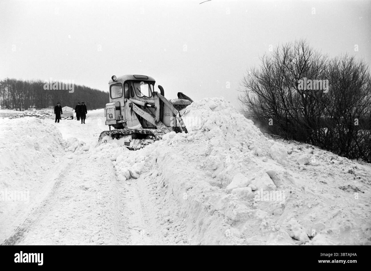 Autobus N.Z.H. con ghiaccio a De Zilk Sassenheim Ruigenhoek ecc. Noord Zuid Hollandse Vervoersmaatschappij N.V. NZ, Whizgle News, Dutch Desk, Paesi Bassi, 1950 - 2000 il 02-01-1963. L'immagine mostra questi argomenti. La scena presenta una vasta distesa di terreno carico di neve, con torreggianti mucchi di neve bianca che creano un netto contrasto contro il cielo grigio. Uno spazzaneve per impieghi gravosi occupa il centro della scena, con il suo telaio in metallo e le lame rivestite in strati di ghiaccio e neve, a indicare la recente attività di sgombero del percorso. Questo macchinario ha un aspetto piuttosto industriale, grazie alla sua struttura robusta e al design pratico Foto Stock