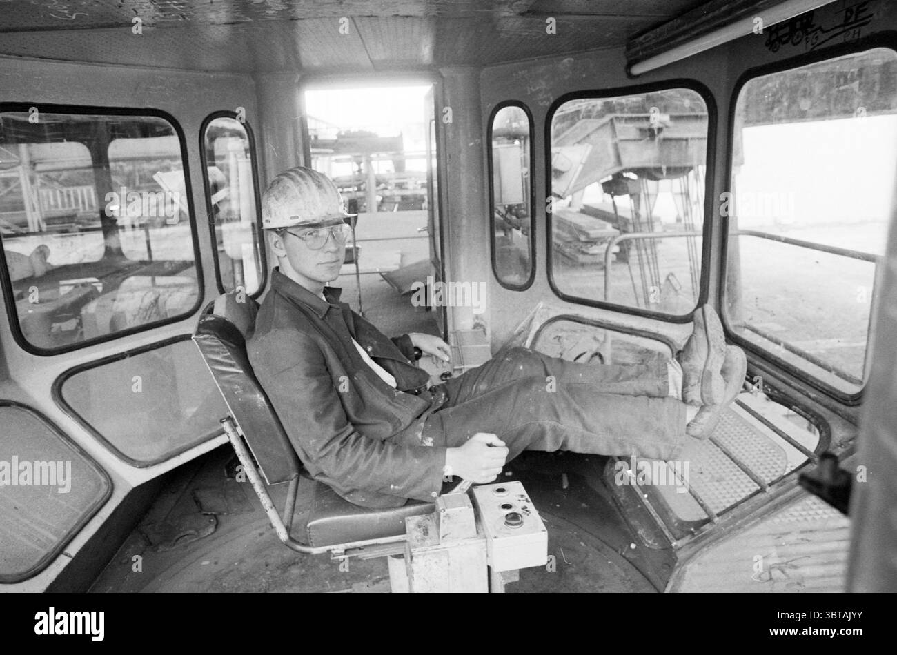 Scuola di demolizione Kerkstraat IJmuiden cantieri di demolizione IJmuiden Kerkstraat Paesi Bassi, Whizgle News, Dutch Desk, Paesi Bassi, 1950 - 2000 il 16-07-1986. L'immagine include questi argomenti. In una scena prevalentemente monocromatica, un giovane uomo siede in una cabina operatore chiusa, che ricorda un ambiente industriale. Gli interni della cabina sono caratterizzati da superfici metalliche, con un aspetto resistente agli agenti atmosferici che indica un uso intensivo. Le pareti sono rivestite da pesanti pannelli di vetro, che consentono una vista ampia dell'ambiente circostante pur essendo un po' nebbiose o sporche, suggerendo un ambiente di lavoro Foto Stock