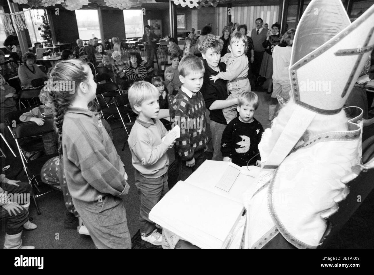 Sinterklaas Afternoon Haarlems Dagblad ecc. e IJmuider Courant e Koeri, Whizgle News, Dutch Desk, Paesi Bassi, 1950 - 2000 il 03-12-1986. L'immagine contiene questi argomenti. La scena è ambientata in un vivace spazio interno, probabilmente una comunità o un centro di attività, caratterizzato da un'atmosfera calda e invitante. In primo piano, un gruppo di bambini si trova in un semicerchio, le loro espressioni un mix di curiosità ed eccitazione. Ogni bambino ha un aspetto unico, indossando una varietà di maglioni e abbigliamento casual, mostrando una miscela di colori, tra cui tonalità luminose e motivi giocosi come stelle e stri Foto Stock