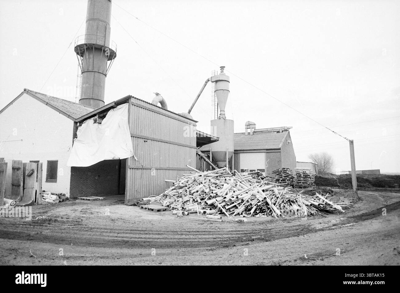 Esterno della fabbrica di tubi in PVC., Whizgle News, Dutch Desk, Paesi Bassi, 1950 - 2000. L'immagine mostra questi argomenti. La scena raffigura un'area industriale caratterizzata da una giustapposizione di strutture e materiali. In primo piano, una grande pila di rottami di legno e detriti viene accumulata sul terreno, suggerendo un'attività di costruzione o demolizione in corso. Questi resti sono di varie dimensioni, alcuni ritorti e rotti, indicando che hanno visto giorni migliori. A destra si può osservare un insieme di edifici, principalmente costruiti in metallo ondulato e legno intemprato, che accennano alla loro età Foto Stock