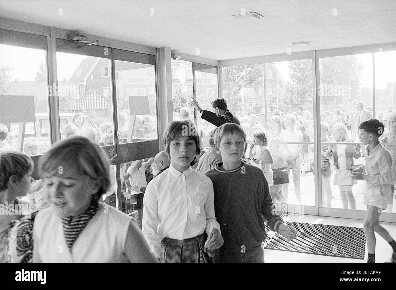 1st clienti Stuif Stuif Stuif Stuif, Whizgle News, Dutch Desk, Paesi Bassi, 1950 - 2000 il 14-07-1969. Questi argomenti sono mostrati nell'immagine. La scena cattura un momento in un ambiente vivace, probabilmente in una scuola o in un centro comunitario. Si vede un gruppo di bambini, con due ragazzi in primo piano. Il ragazzo sulla sinistra indossa una camicia bianca button-up, mentre quello accanto a lui è vestito con un maglione scuro e pantaloncini di colore chiaro. Le loro espressioni suggeriscono curiosità o eccitazione mentre attraversano l'ingresso. L'impostazione è dotata di grandi sportelli in vetro che si aprono su ot Foto Stock
