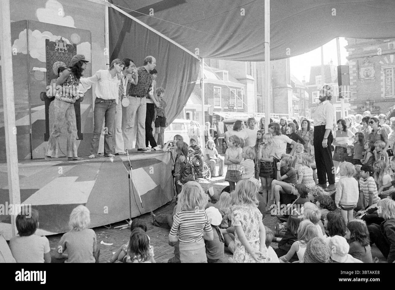 Teatro di strada Grote Markt Theater Haarlem Grote Markt Paesi Bassi, Whizgle News, Dutch Desk, Paesi Bassi, 1950 - 2000 il 10-08-1973. Questi argomenti vengono visualizzati nell'immagine. In un ambiente all'aperto vivace, una vivace performance si svolge sotto un grande baldacchino. Il palco, adornato da uno sfondo dipinto di nuvole e colline, presenta diversi artisti impegnati in una scena animata. Gli individui vestiti con un mix di abbigliamento colorato e informale mostrano una gamma di espressioni, dalla gioia alla sorpresa, suggerendo una narrazione comica o giocosa. In primo piano, un pubblico diversificato, composto principalmente da Foto Stock