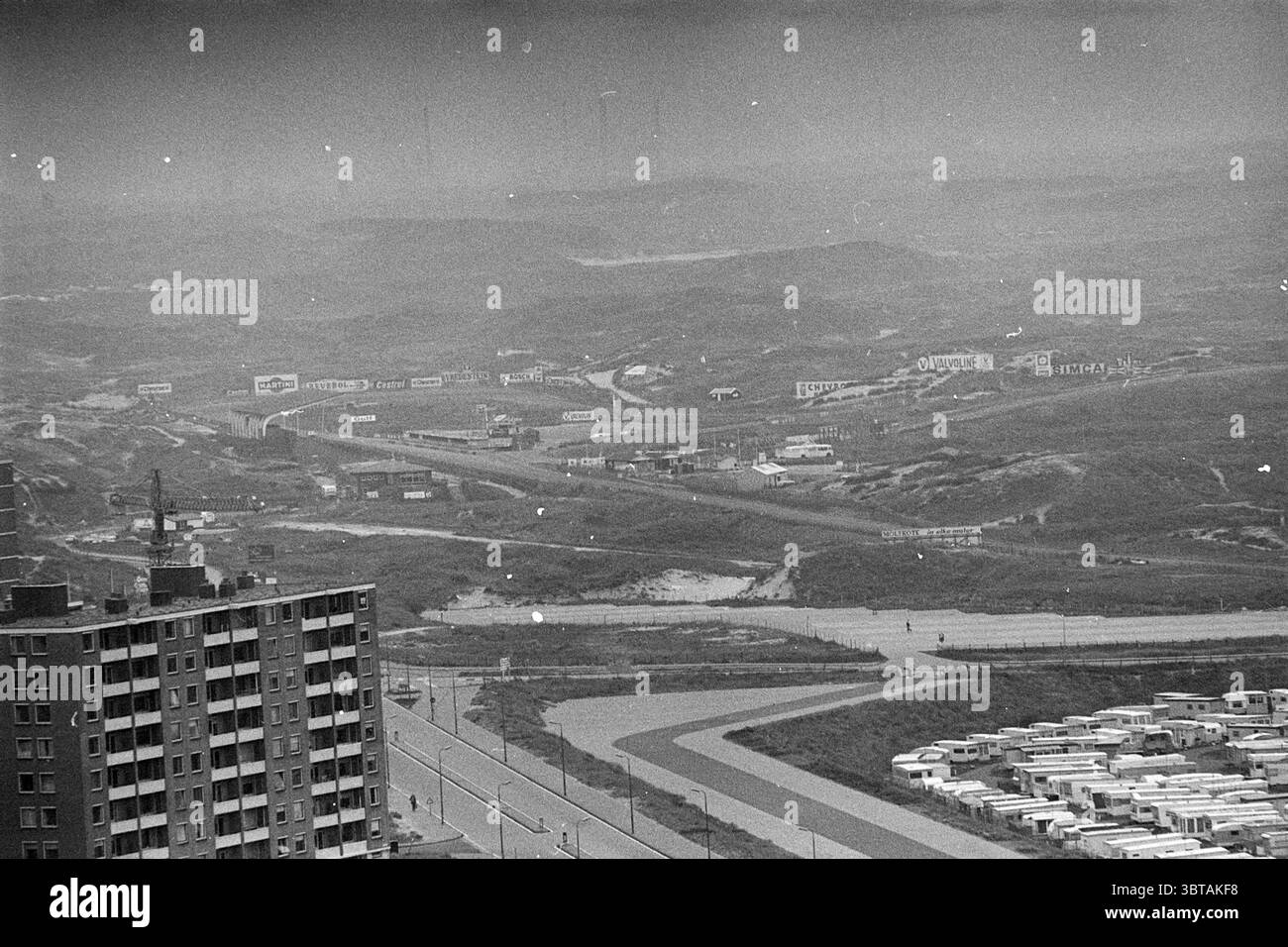 Circuito Zandvoort Overviews, Whizgle News, Dutch Desk, Paesi Bassi, 1950 - 2000 on 29-09-1972. L'immagine include questi argomenti. La scena presenta una vista estesa di un paesaggio in qualche modo industrializzato, bagnato da una scala di grigi silenziosa che gli conferisce un'atmosfera nostalgica o malinconica. In primo piano, un alto edificio a blocchi con finestre multiple si erge come una struttura solida, la sua facciata segnata da linee angolari e un modello di elementi ripetuti. Oltre l'edificio, il terreno si estende, dominato da colline ondulate e macchie di terra che appaiono aride, suggerendo un'area che m Foto Stock