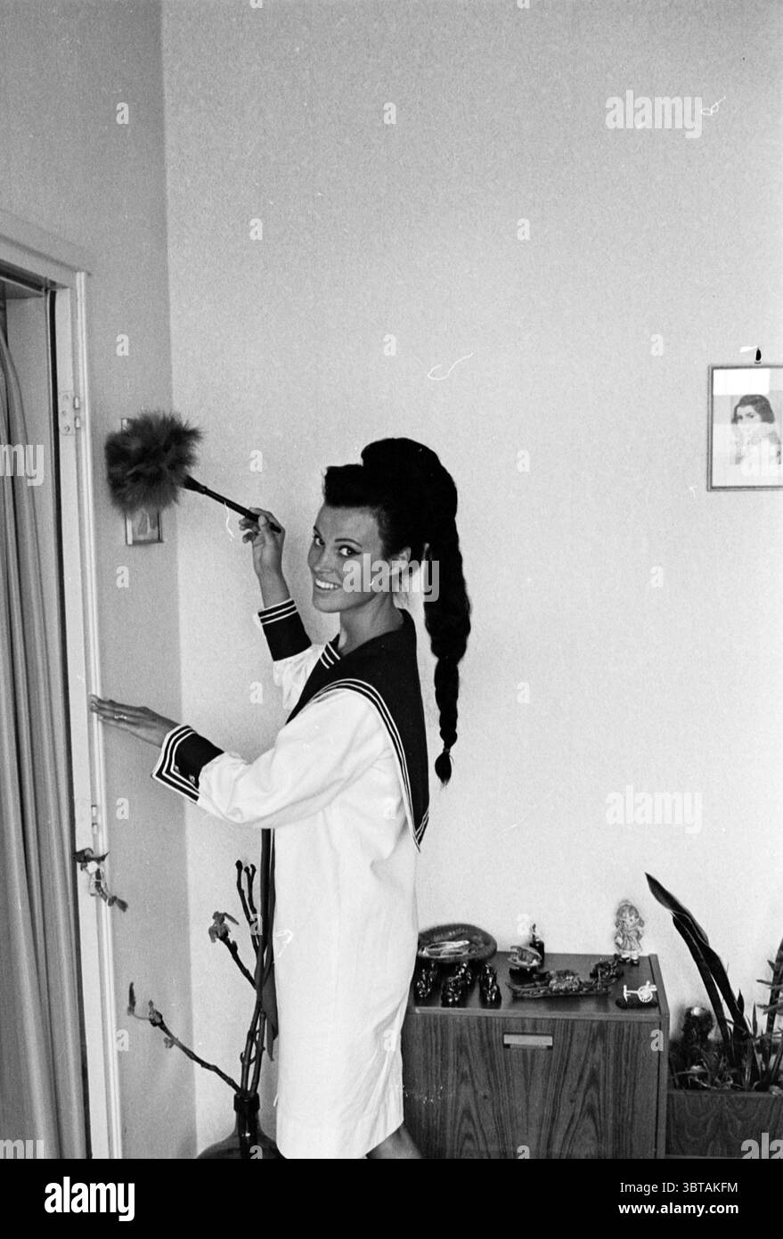 Mannequin Marianna v.d. Heyden Models, Whizgle News, Dutch Desk, Paesi Bassi, 1950 - 2000 il 04-06-1963. Questi argomenti vengono visualizzati nell'immagine. In un ambiente interno luminoso, una donna si erge allegramente, impegnandosi in un lavoro. Indossa un abito di colore chiaro con un particolare collare a vela, decorato con strisce scure, che aggiunge un tocco di giocosità al suo look. I suoi capelli hanno un aspetto alto e voluminoso, simile a un tocco drammatico, che le conferisce un aspetto classico ma vivace. La camera presenta pareti dai colori neutri che completano l'arredo minimalista. Un tavolino in legno è posizionato nelle vicinanze, Foto Stock