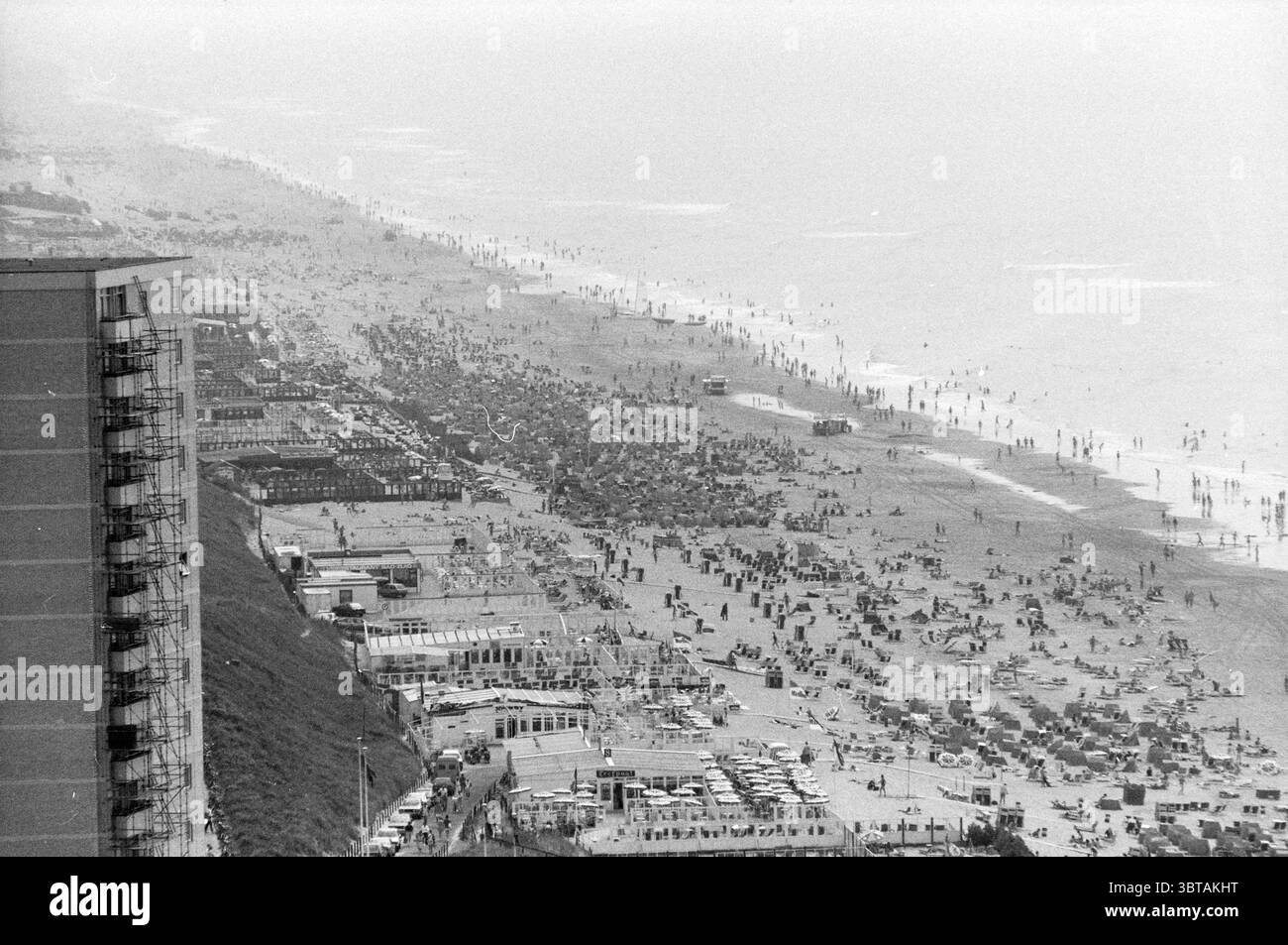 Panorama Zandvoort da Bouwes Overviews Zandvoort Zandvoort, Whizgle News, Dutch Desk, Paesi Bassi, 1950 - 2000 il 23-06-1986. Questi sono gli argomenti nell'immagine. La scena si snoda lungo una vivace spiaggia, caratterizzata da numerose persone che si godono una giornata di sole sull'acqua. Da un lato, si erge un alto edificio che getta una lunga ombra sulla distesa di sabbia sottostante. La spiaggia è densamente popolata, con gruppi di ombrelloni e sedie a sdraio che creano un arazzo di colori contro la sabbia beige. In primo piano, gruppi di bagnanti si rilassano sugli asciugamani, alcuni si sono impegnati a conversare mentre si trovava Foto Stock