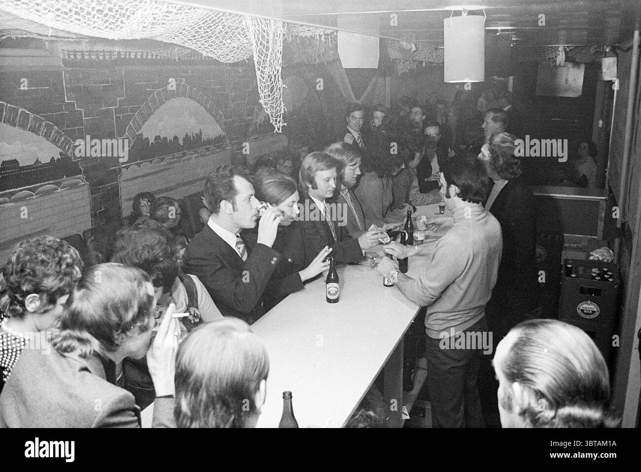 Gruppo di persone in un bar durante una festa., Whizgle News, Dutch Desk, Paesi Bassi, 1950 - 2000. L'immagine mostra questi argomenti. La scena si svolge all'interno di un locale poco illuminato che trasuda un'atmosfera vintage. L'attenzione è su un bar lungo e stretto, dove si riuniscono un gruppo di clienti. Su un lato del bar, diversi individui sono impegnati in una conversazione, alcuni che tengono bevande mentre altri si appoggiano casualmente al bancone. La superficie del bar e' piena di varie bottiglie di bevande, a indicare un'atmosfera vivace. I clienti sono vestiti in stili tipici dell'epoca, con abiti e abiti, a Foto Stock