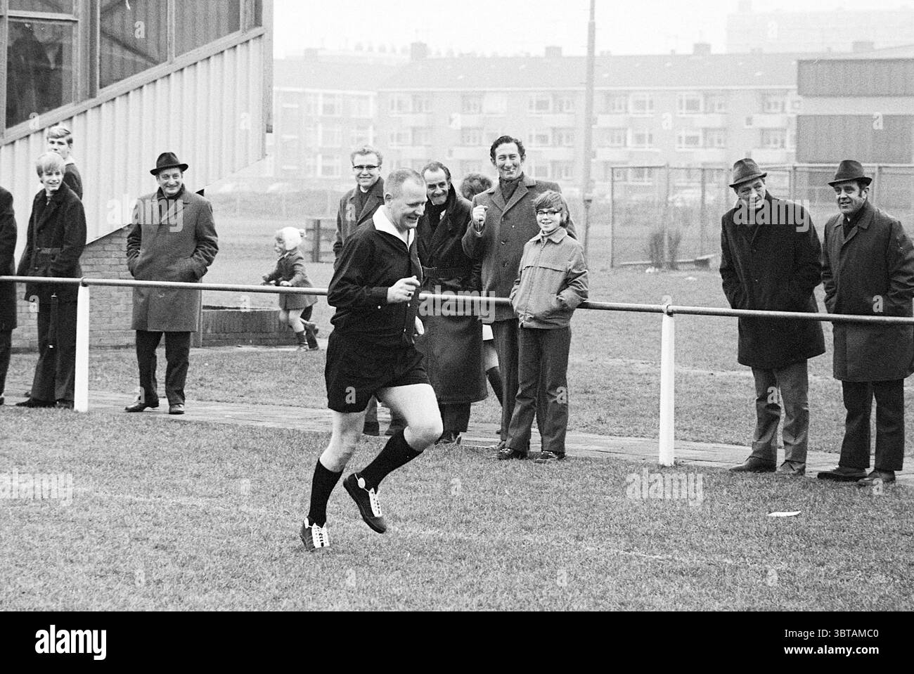 Stormvogels - Red White Football V.S.V. Velsen Stormvogels, Whizgle News, Dutch Desk, Paesi Bassi, 1950 - 2000 il 07-02-1971. L'immagine include questi argomenti. La scena raffigura un dinamico evento sportivo all'aperto, probabilmente una partita di calcio, che si svolge su un campo erboso. Un arbitro, vestito con un'uniforme nera con pantaloncini corti e un fischio, si vede volare attraverso il campo. La sua espressione suggerisce determinazione, e contrasta nettamente con gli spettatori circostanti. La folla è posizionata lungo una recinzione, mostrando un mix di reazioni. Molti uomini sono vestiti con cappotti e cappelli scuri e formali, indic Foto Stock