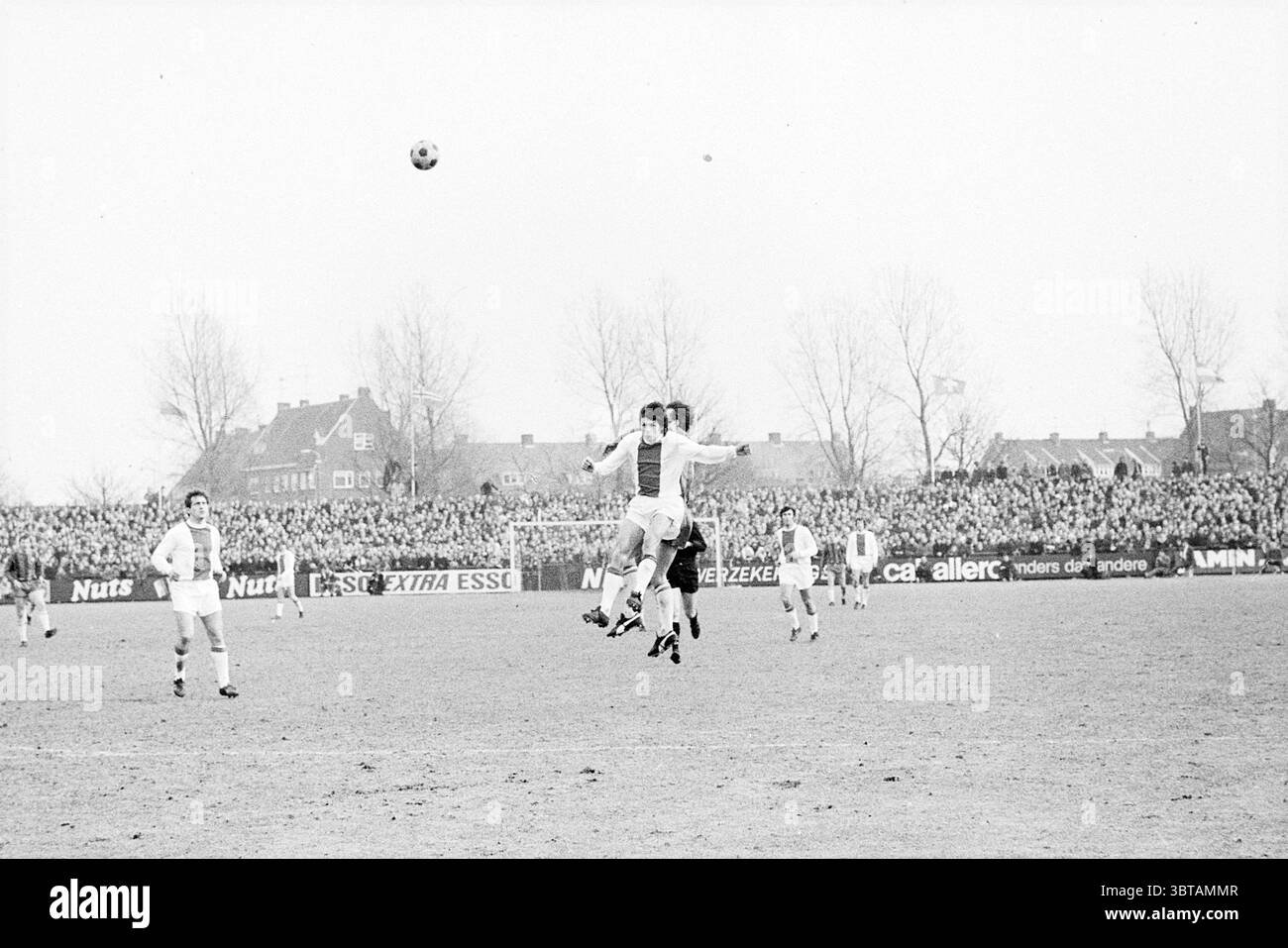 Haarlem - Ajax Football Haarlem, Whizgle News, Dutch Desk, Paesi Bassi, 1950 - 2000 il 08-03-1970. L'immagine mostra questi argomenti. La scena cattura un momento dinamico in una partita di calcio che si svolge all'aperto in una giornata grigia. In primo piano, due giocatori stanno saltando in aria, braccia allungate, mentre lottano per il controllo della palla. Entrambi gli atleti sono vestiti con divise da calcio classiche, con una maglia bianca e l'altra con una combinazione di rosso e bianco. Lo sfondo è pieno di una grande folla di spettatori, i loro volti oscurati ma la massa di corpi che suggeriscono eccitazione Foto Stock