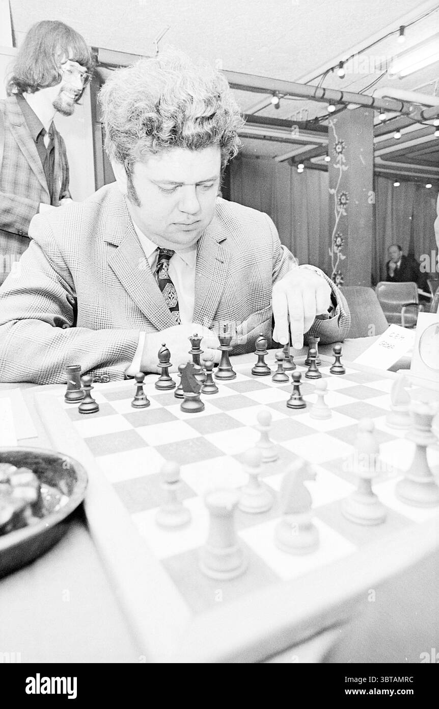 Evento inaugurale Blast Furnace Chess Tournament, Whizgle News, Dutch Desk, Paesi Bassi, 1950 - 2000 il 15-01-1972. L'immagine contiene questi argomenti. In un ambiente interno vivace, un uomo concentrato è seduto a un tavolo da scacchi, profondamente impegnato in una partita. Sfoggia una tuta testurizzata di colore chiaro che contrasta con le ricche sfumature scure dei pezzi di legno degli scacchi sulla scacchiera davanti a lui. I suoi capelli sono ricci e voluminosi, aggiungendo un fascino vintage al suo aspetto. Si piega leggermente in avanti, la sua mano è appoggiata su una pedina nera, contemplando la sua prossima mossa. La scacchiera stessa è un classico a scacchi Foto Stock