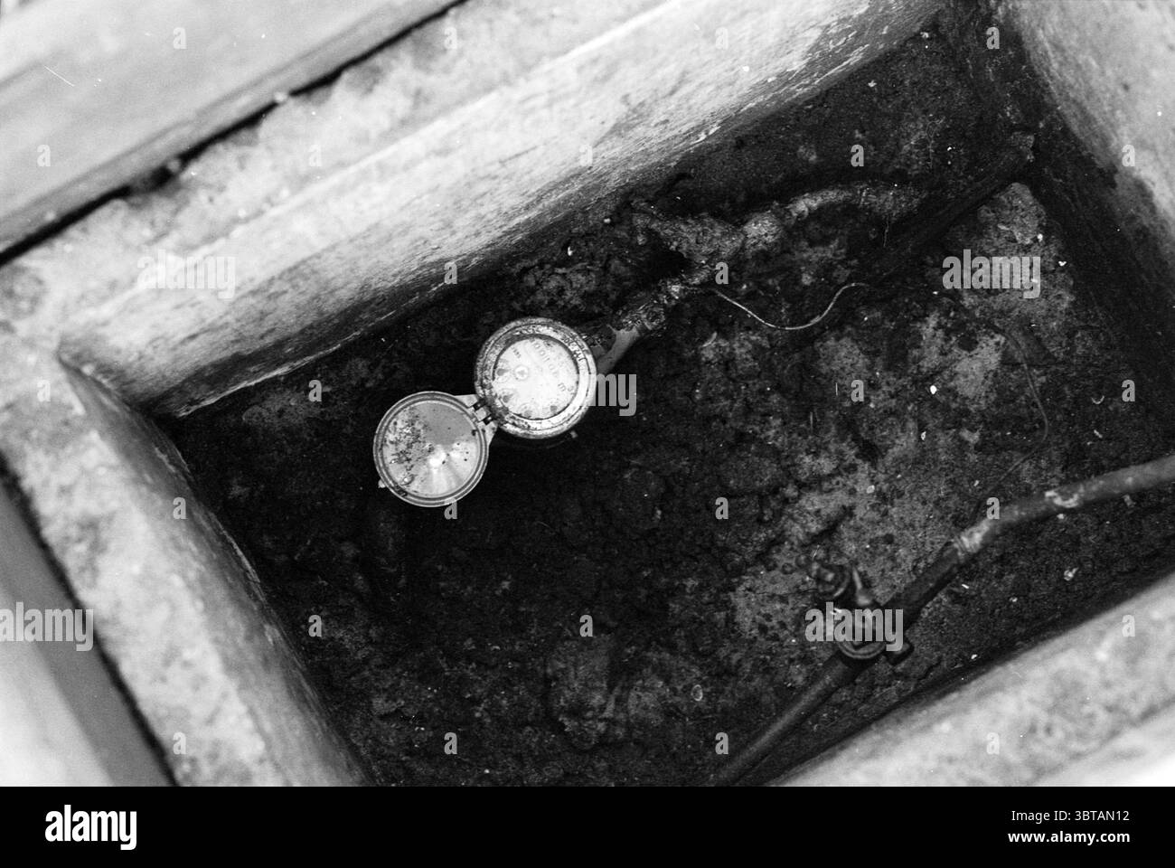 Contatore d'acqua, Whizgle News, Dutch Desk, Paesi Bassi, 1950 - 2000 il 23-07-1992. Questi sono gli argomenti nell'immagine. In uno spazio sotterraneo scarsamente illuminato, è visibile una cavità rettangolare, riempita di terreno scuro e ricco che suggerisce che non è stato disturbato per qualche tempo. Al centro di questa scena, due strumenti circolari sono parzialmente esposti e le loro superfici metalliche riflettono deboli sbiadimenti di luce. I manometri hanno un aspetto obsoleto, con segni di usura che suggeriscono un lungo utilizzo nella misurazione del flusso o della pressione. Intorno agli strumenti, i tubi sottili e aggrovigliati si estendono dai lati di t Foto Stock