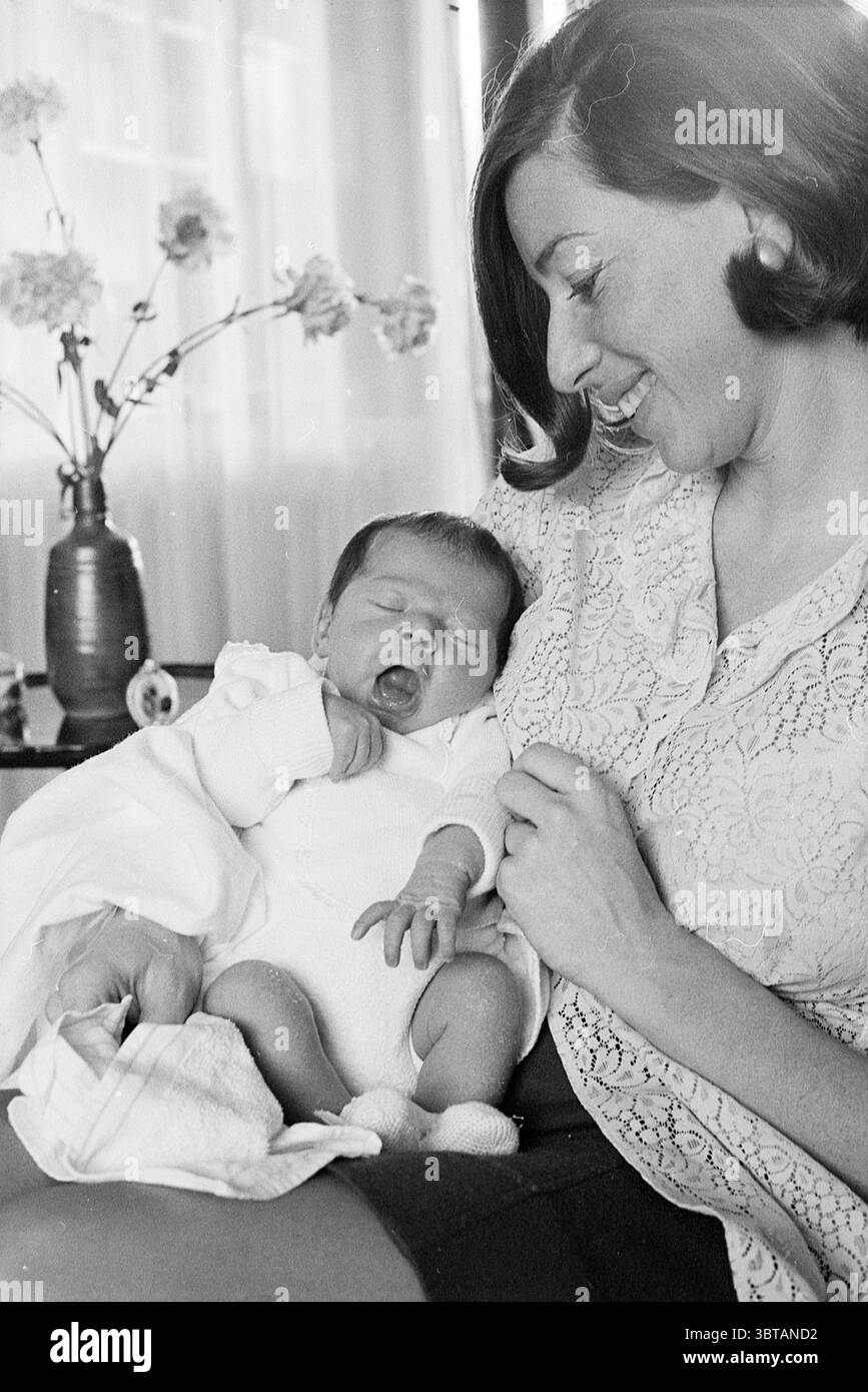 Madre con bambino, Whizgle News, Dutch Desk, Paesi Bassi, 1950 - 2000 il 02-1969. Questi argomenti vengono visualizzati nell'immagine. In un ambiente interno accogliente, una donna è seduta comodamente, cullando un bambino addormentato tra le braccia. La composizione si concentra sulla loro intima connessione. La donna, con i capelli scuri a spalla, indossa un capo dai colori chiari e fantasia che aggiunge un tocco morbido alla scena. La sua espressione trasmette calore e gioia, accentuata da un sorriso gentile mentre guarda il bambino. Il bambino, vestito con un semplice vestito bianco, appare tranquillo, con gli occhi chiusi e un sottile sbadiglio, Foto Stock