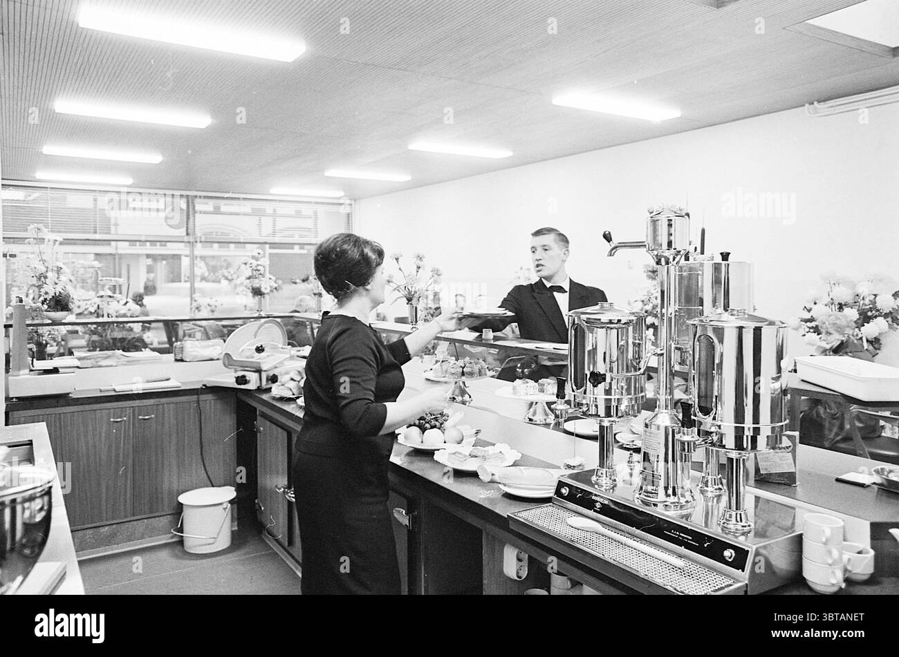 Tempelierstraat stop building Interior North South Hollandse Vervoersmaatschappij N.V. Haarlem Tempeliersstraat Paesi Bassi, Whizgle News, Dutch Desk, Paesi Bassi, 1950 - 2000 su 11-11-1963. Questi argomenti vengono visualizzati nell'immagine. La scena si sviluppa in un ambiente luminoso e moderno, caratterizzato da un'estetica pulita e minimalista. Le camere sono prevalentemente bianche, con caldi accenti di legno che suggeriscono un'atmosfera accogliente. In primo piano, una donna che indossa un elegante abito nero è impegnata in azione, il suo corpo leggermente girato mentre consegna una tazza a un uomo in piedi dietro una macchiatura lucida Foto Stock