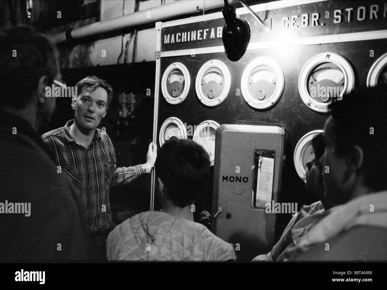 Interno di una stazione di pompaggio del vapore., Whizgle News, Dutch Desk, Paesi Bassi, 1950 - 2000. L'immagine include questi argomenti. La scena è ambientata in un ambiente scarsamente illuminato, con una forte focalizzazione su un pannello di controllo centrale ornato da diversi indicatori circolari. Ogni indicatore visualizza varie letture, suggerendo il funzionamento complesso dei macchinari. Il pannello di controllo ha una lucentezza metallica ed è incorniciato da un design semplice e pratico. Un gruppo di quattro persone è impegnato in una conversazione nelle vicinanze. Un uomo sta in piedi in modo prominente davanti al pannello, spiegando animatamente qualcosa, la sua espressione sul serio e fo Foto Stock
