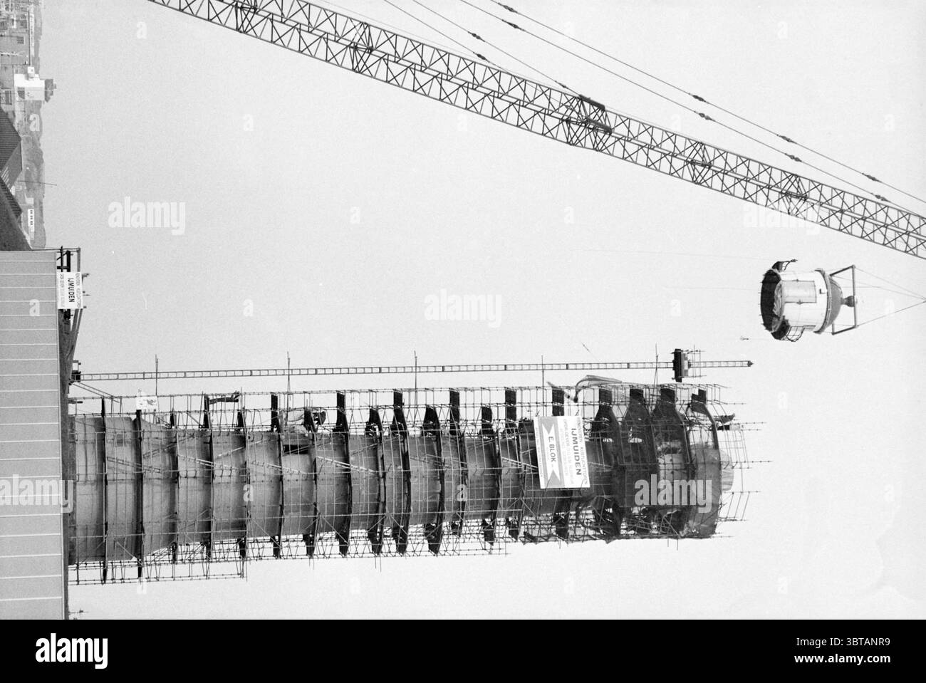 Rinnova faro faro IJmuiden Faro IJmuiden Paesi Bassi, Whizgle News, Dutch Desk, Paesi Bassi, 1950 - 2000 il 19-05-1987. L'immagine include questi argomenti. La scena cattura un grande cantiere dominato da un'alta struttura cilindrica parzialmente completata. La torre è racchiusa in impalcature, con un aspetto a rete con un reticolo di travi metalliche che attraversano l'esterno. La struttura si innalza significativamente nel cielo, suggerendo che sarà un alto edificio. A sinistra, una gru sporgente si estende dal suolo, con il suo lungo braccio che si estende fino a. Foto Stock