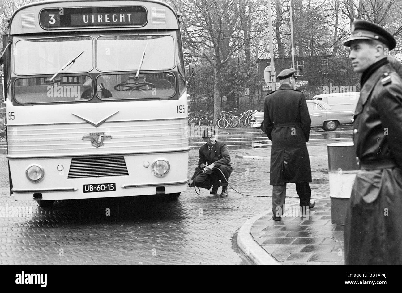 Sciopero degli autobus di Riots Kennemerplein Strikes, Whizgle News, Dutch Desk, Paesi Bassi, 1950 - 2000 il 15-11-1963. L'immagine include questi argomenti. In una vivace scena urbana, un autobus d'epoca domina il primo piano, mostrando un design distintivo tipico dei trasporti pubblici della metà del XX secolo. L'autobus e' dipinto in un classico bianco e adornato con linee eleganti. Il suo cartello di destinazione recita "UTRECHT", che indica chiaramente il suo percorso, mentre il numero "3" esposto in alto aggiunge al senso di ordine tipico di un sistema di transito cittadino. Accanto all'autobus, un uomo con un'uniforme scura si inginocchia accanto al vehi Foto Stock