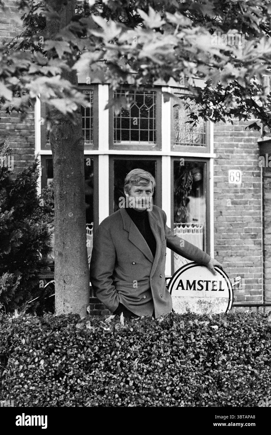 Bert Voskuil book, Whizgle News, Dutch Desk, Paesi Bassi, 1950 - 2000 il 03-10-1994. Questi sono gli argomenti nell'immagine. In un ambiente tranquillo e urbano, un uomo si trova casualmente contro una siepe ben rifilata, incorniciando la sua figura nel verde. Il suo abbigliamento è formale ma rilassato, con un cappotto doppiopetto che contrasta nettamente con le morbide texture del fogliame circostante. L'uomo ha un'espressione premurosa, il suo sguardo si è leggermente spostato di lato, aggiungendo un'aria di contemplazione alla scena. Alla sua sinistra, c'è un assaggio di una bicicletta vintage, che presta una qualità nostalgica. Il Foto Stock