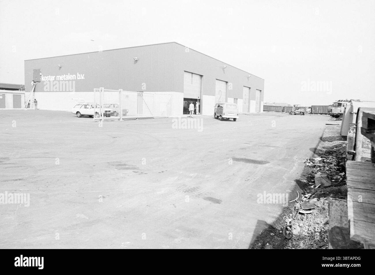 Esterno Koster Metals Beverwijk Exterior Metal Beverwijk Randweg Paesi Bassi, Whizgle News, Dutch Desk, Paesi Bassi, 1950 - 2000 il 02-09-1983. L'immagine include questi argomenti. La scena è caratterizzata da un grande edificio industriale caratterizzato da un rivestimento in metallo, dipinto in una tonalità fresca e silenziosa che contrasta con l'ambiente circostante. La struttura dispone di diverse ampie porte del vano di carico, parzialmente aperte, che suggeriscono attività all'interno. In primo piano si dispiega un'ampia area pavimentata, principalmente in cemento, con pochi veicoli parcheggiati sparsi in tutto. Due auto sono visibili, una a Foto Stock