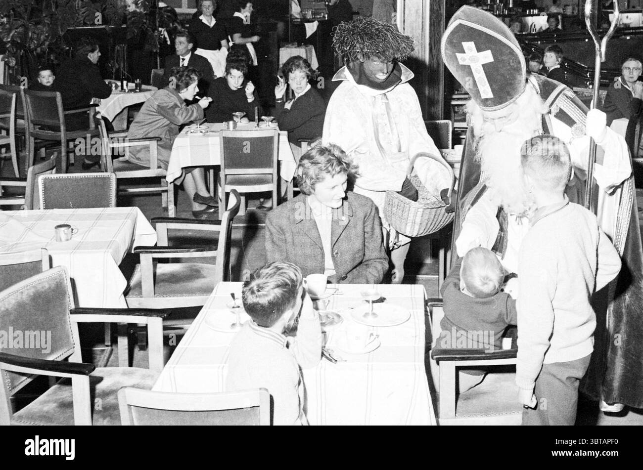 Saint Nicholas at Vroom and Dreesmann Lunchroom St. Nicholas Sinterklaas, Whizgle News, Dutch Desk, Paesi Bassi, 1950 - 2000 il 18-11-1963. L'immagine include questi argomenti. In un vivace ambiente interno, l'atmosfera è piena di calore e senso di comunità. La scena è viva con persone impegnate nella conversazione e che si divertono insieme. I tavoli sono ordinatamente disposti con tovaglie a quadri e diversi gruppi di persone sono seduti, alcuni con tazze e piatti davanti a loro, suggerendo un vivace ambiente da caffetteria o ristorante. In primo piano, una donna con i capelli ricci a Foto Stock