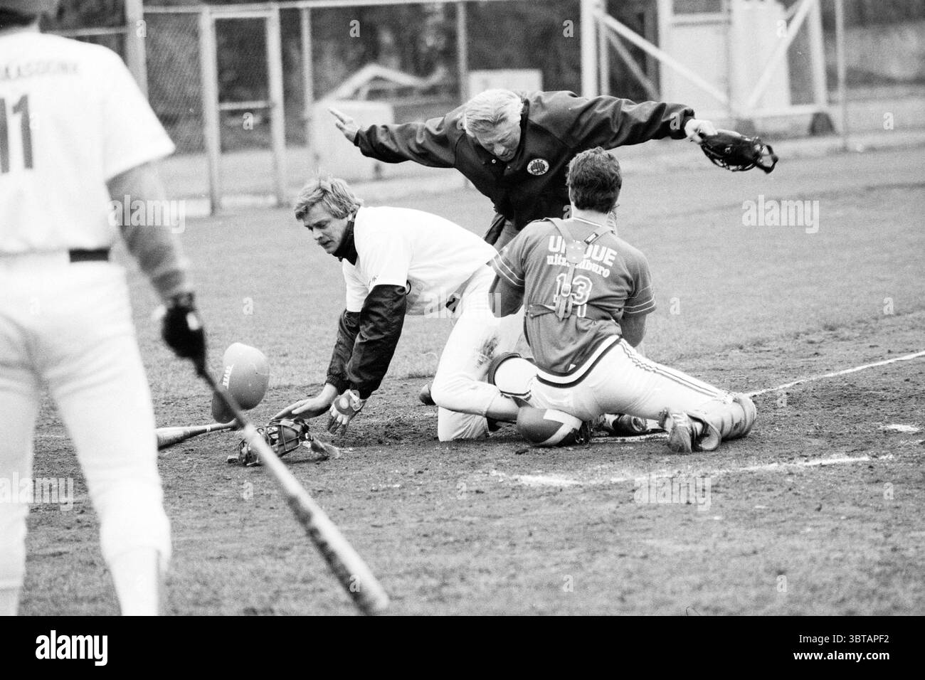 Nicols - Giants Baseball Haarlem Nicols, Whizgle News, Dutch Desk, Paesi Bassi, 1950 - 2000 il 23-04-1989. Questi sono gli argomenti nell'immagine. La scena cattura un momento dinamico su un campo da baseball, presentato in tonalità monocromatiche che amplificano l'intensità dell'azione. Al centro della composizione c'è un giocatore in una posizione accovacciata, vestito con una maglia luminosa contrassegnata con il numero 13, che afferra una mazza da baseball che giace a terra accanto a lui. Sembra essere aggrovigliato in un gioco insolito, con un'espressione concentrata mentre interagisce con gli altri giocatori. Al suo fianco, un altro giocatore in un attimo Foto Stock