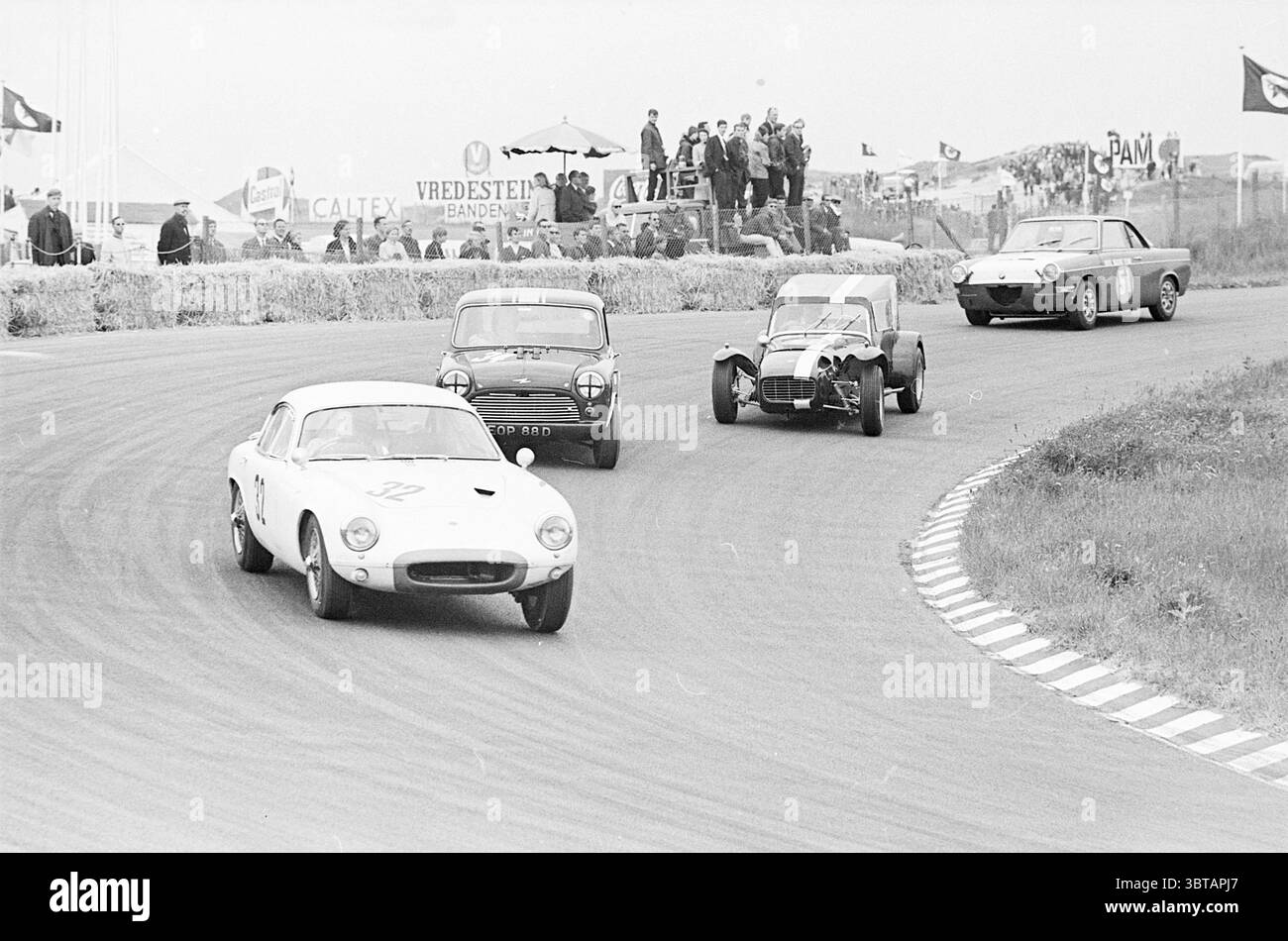 Gare automobilistiche sul circuito di Zandvoort Zandvoort, Whizgle News, Dutch Desk, Paesi Bassi, 1950 - 2000 il 18-06-1967. L'immagine include questi argomenti. La scena cattura un momento emozionante su un circuito, dove le auto d'epoca sono impegnate in una competizione vivace. Il primo piano presenta un'elegante auto sportiva bianca, numerata in modo preminente "12", che fa una curva stretta per navigare lungo la curva del tracciato. Le sue linee lisce e la superficie lucida riflettono i toni tenui dell'ambiente, suggerendo un senso di potenza e velocità. Oltre a questo, due auto classiche più piccole, una scura e l'altra in una tonalità vivace, ap Foto Stock